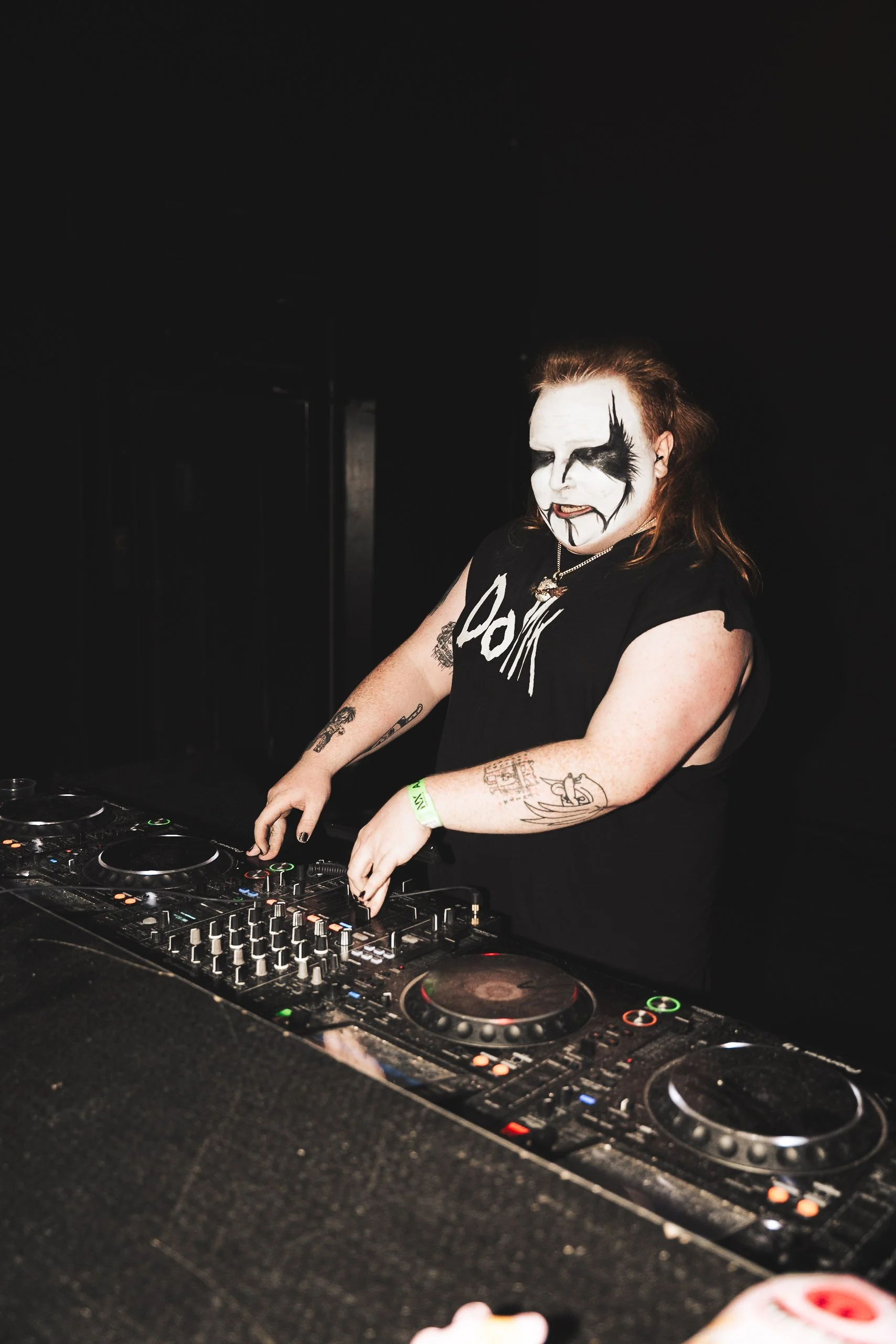 A person with face makeup resembling a black and white clown, wearing a black sleeveless shirt, standing behind DJ equipment, in a dark setting.