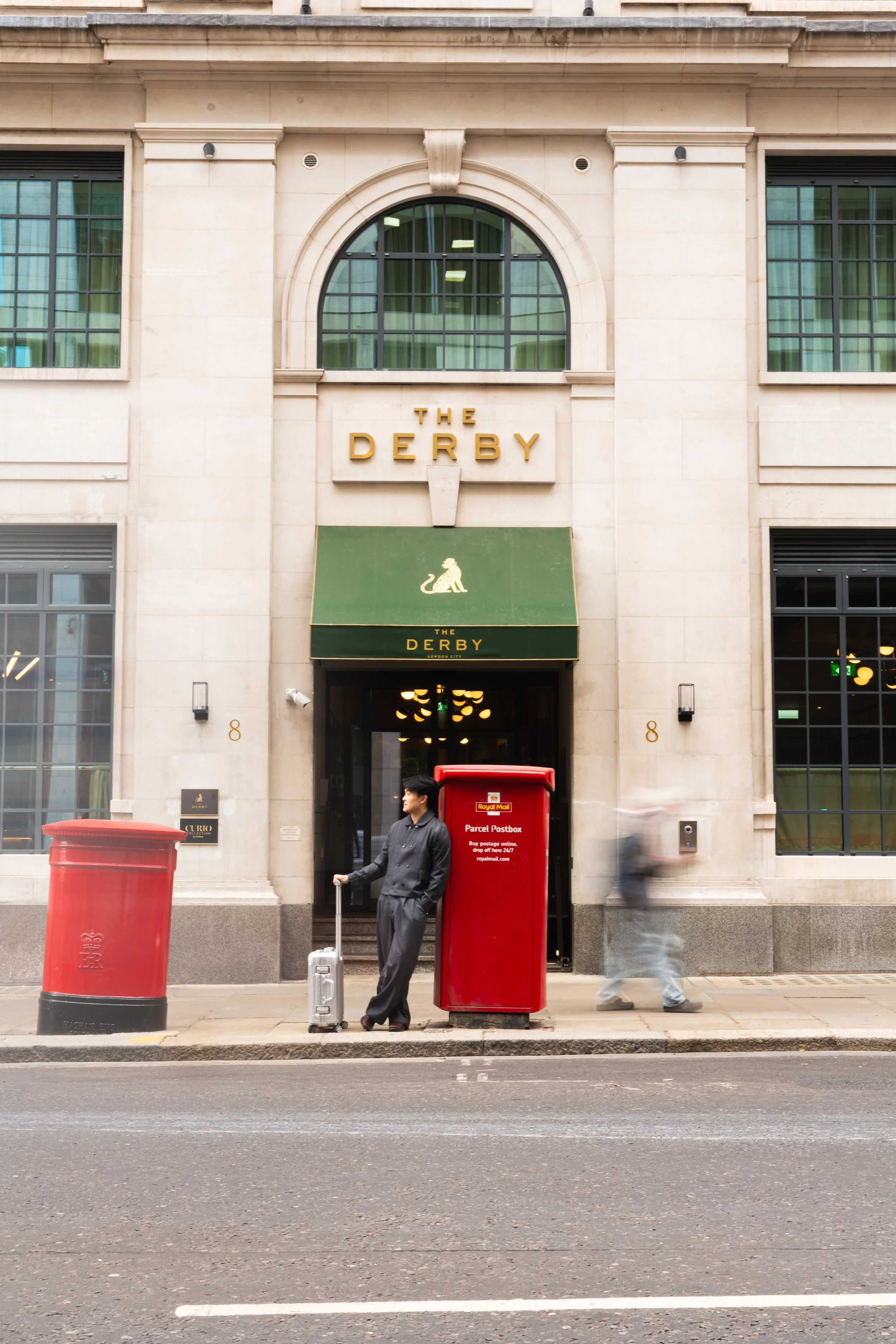 Street view showing two red mailboxes in front of a building labeled "The Derby" with a large arched window. One person with a suitcase is standing near one mailbox, another person is walking past.