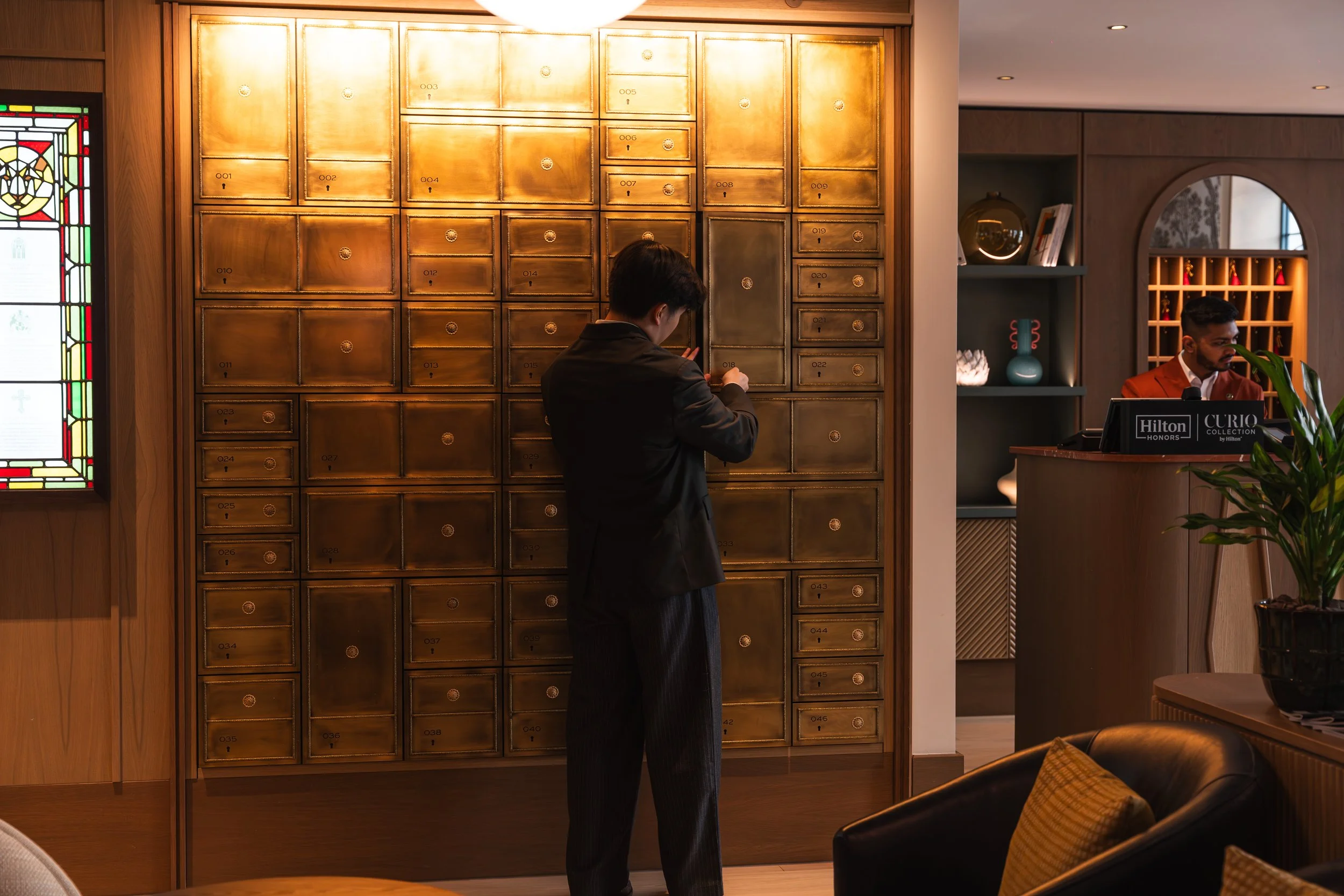 A man in a dark suit stands in front of a wall of small gold-colored lockers, looking at his phone. To the right, a woman with a bun is at a reception desk, and another man with glasses and a reddish-brown blazer stands beside her. The setting is a s
