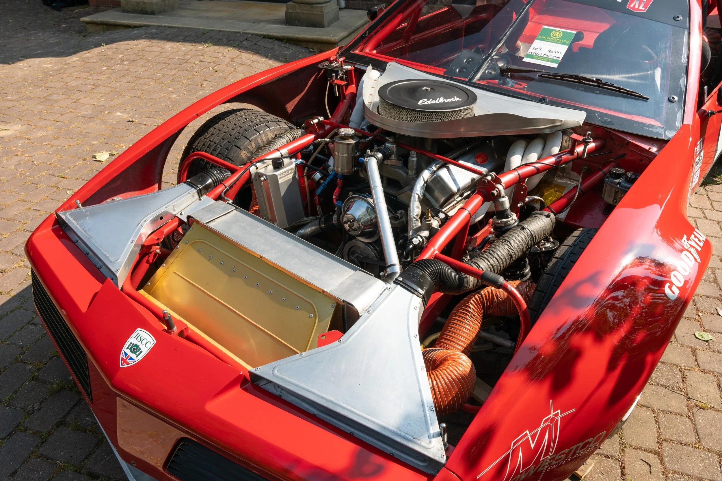 Front view of a vintage red race car with the engine exposed, showing various mechanical components, hoses, and a large Edelbrock air cleaner on top of the engine.