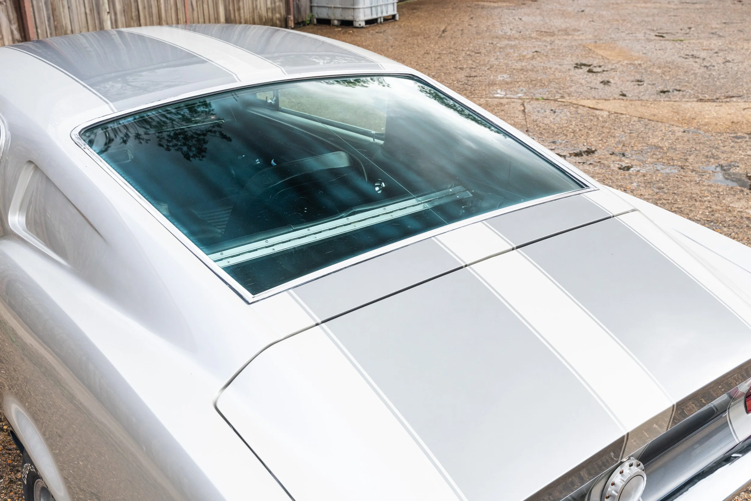 A top view of a silver vintage sports car with a large rear window, showing part of the hood, trunk, and rear wheel on a gravel surface.