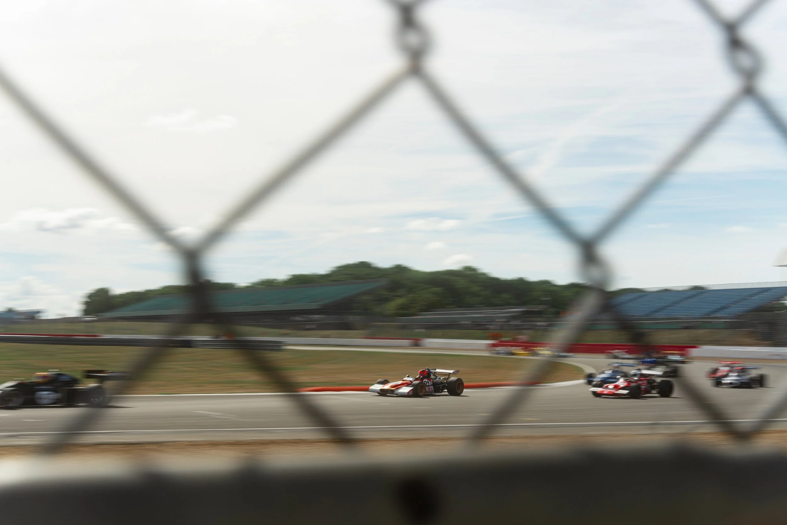 Race cars on a track viewed through a chain-link fence with clouds in the sky.