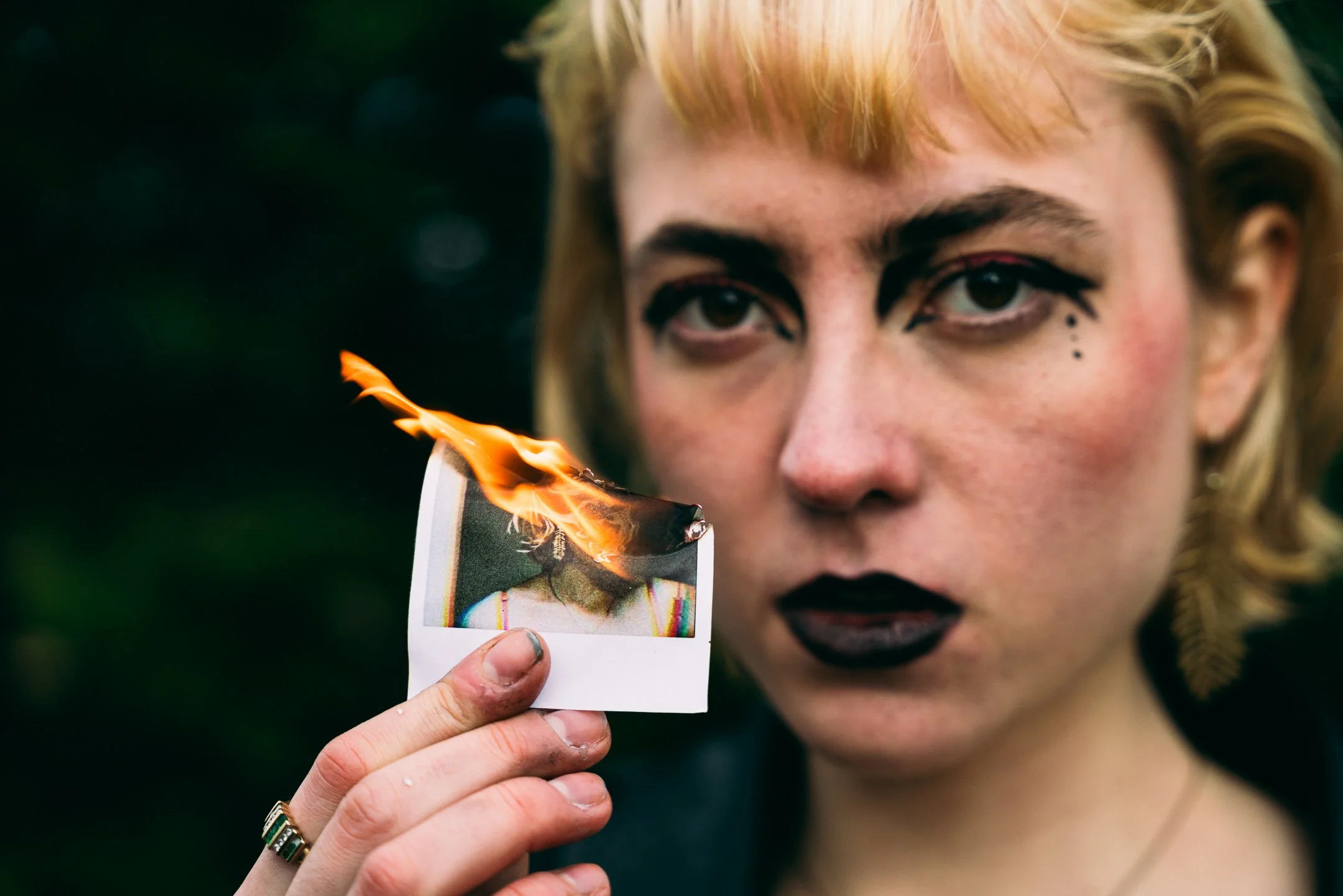 Person with dramatic makeup holding a burning photograph of a woman.