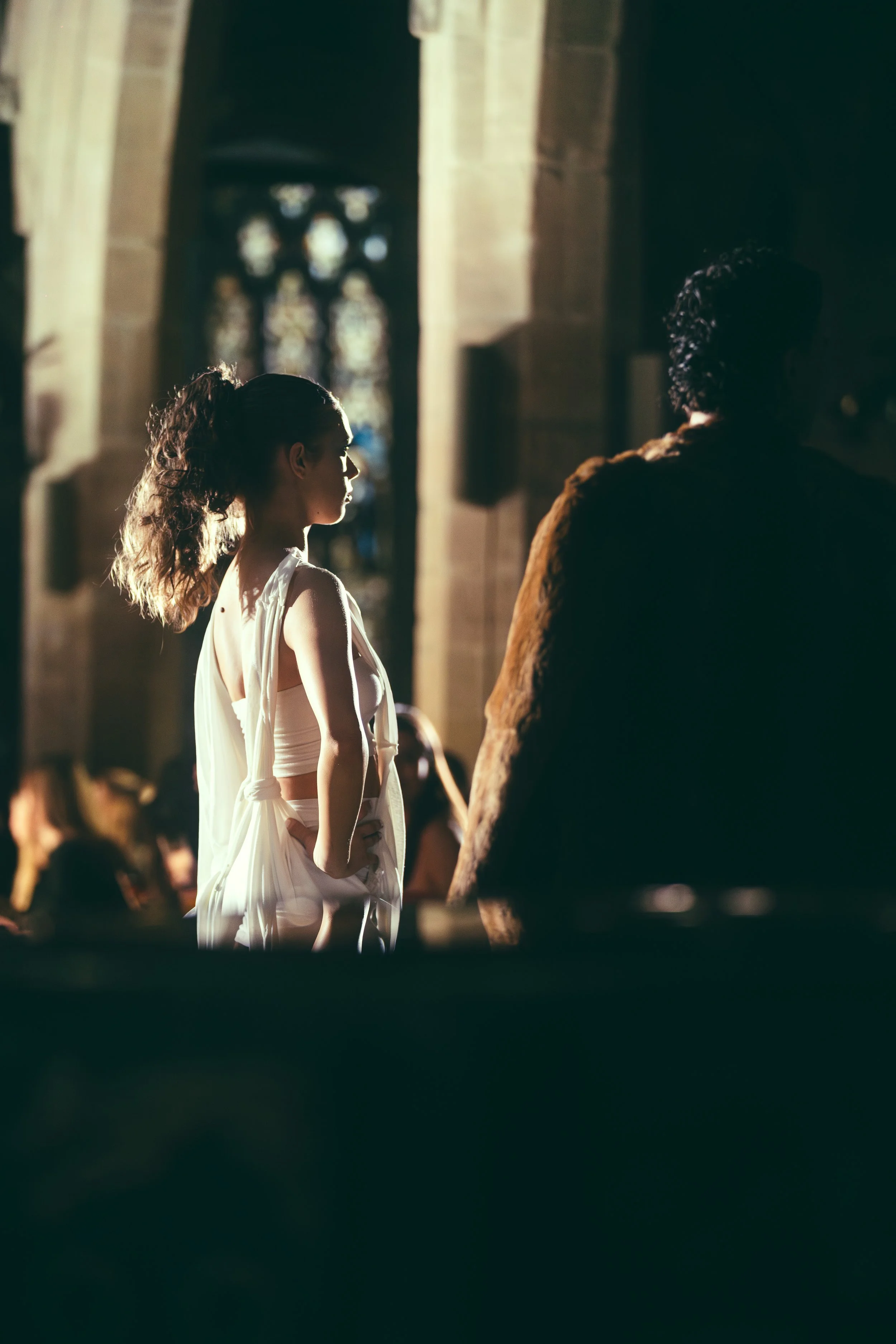 A woman in a white dress standing inside a church or cathedral, facing a man in darker clothing, with stained glass windows in the background.