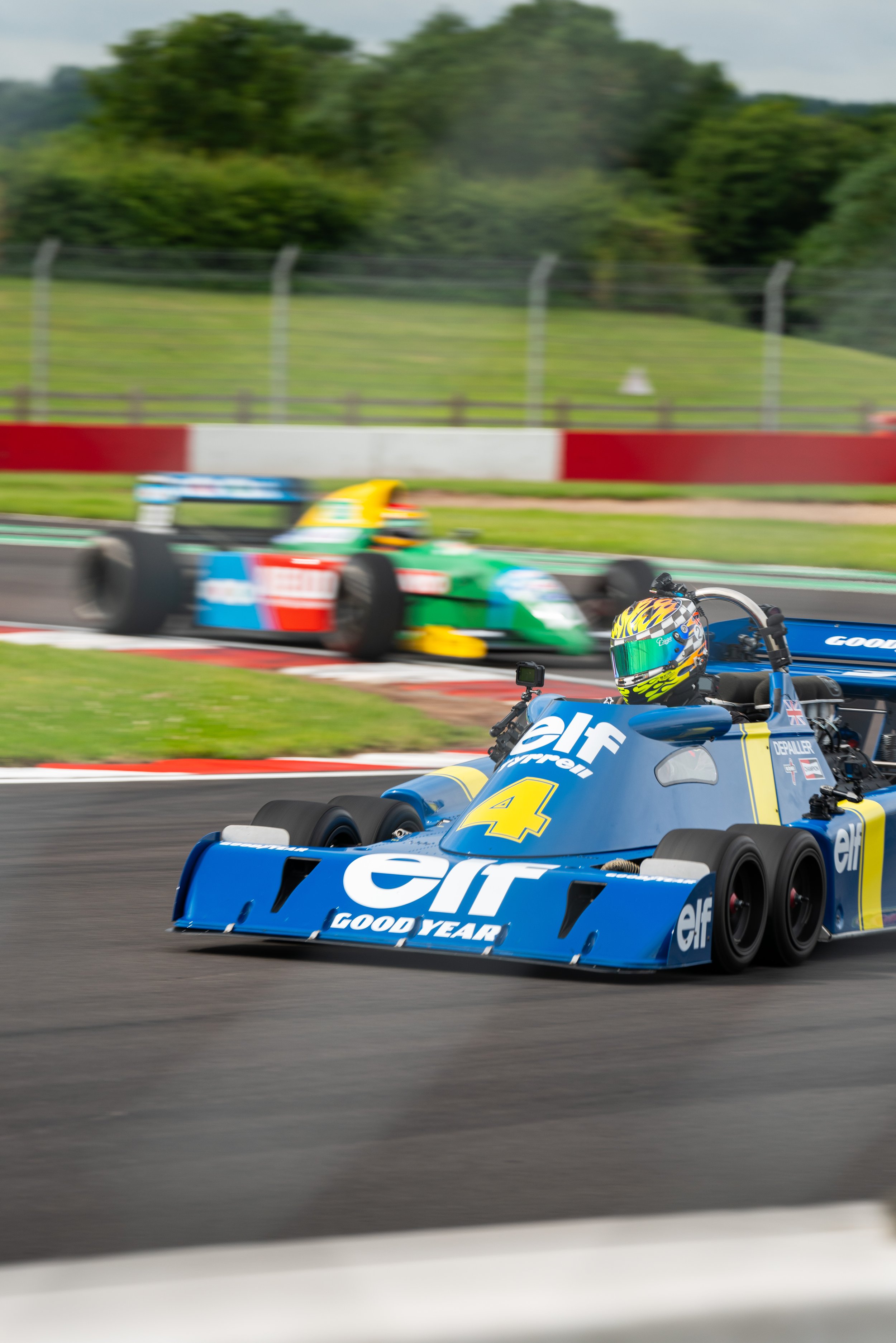 Two race cars on a track, one in blue with ELF and Goodyear branding and the number 4, leading, and a second green and yellow car in the background, with motion blur indicating high speed.