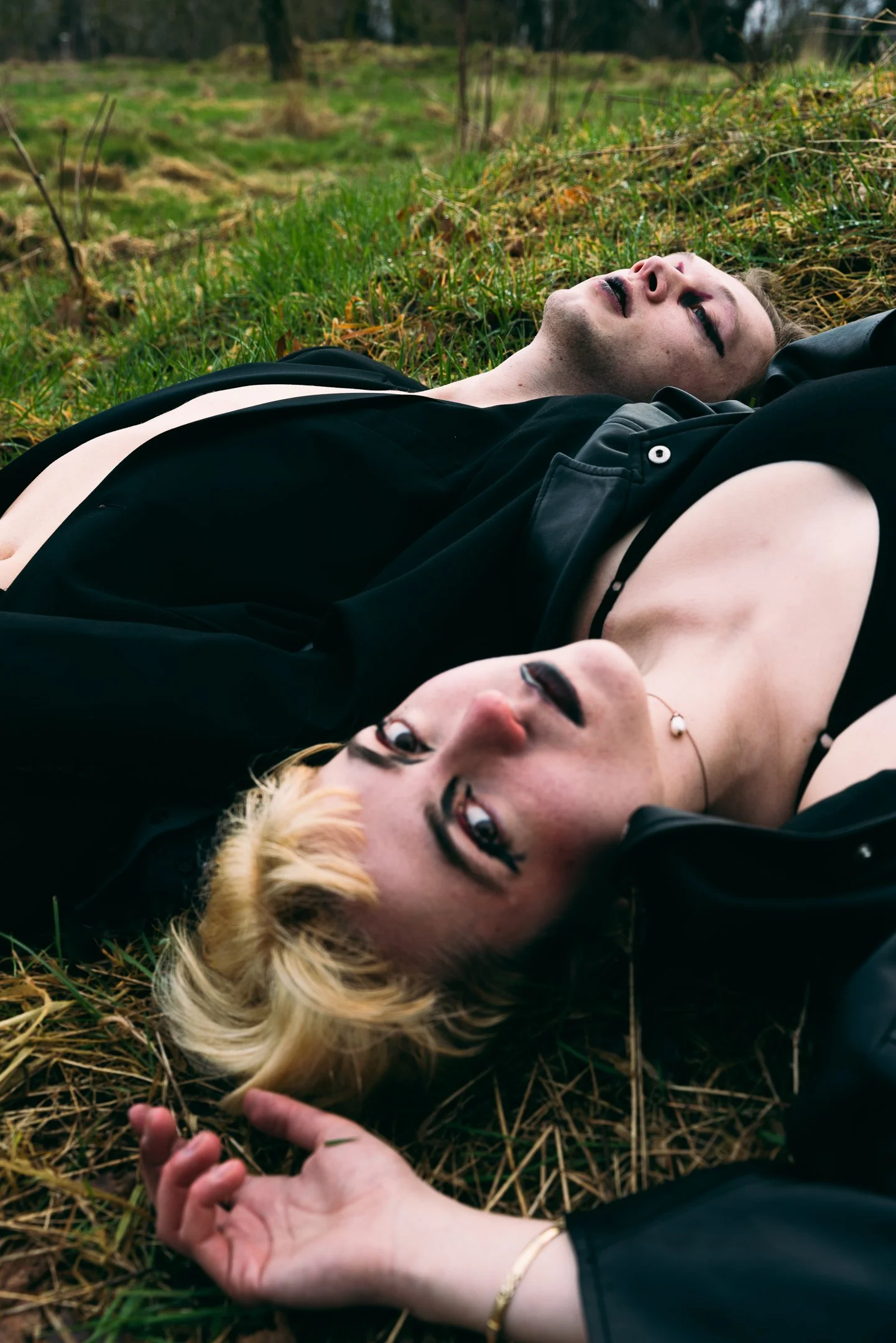 A young woman with blonde hair and dark makeup lying on her back in a field of grass and dried plants, with a young man with dark hair and makeup lying beside her. They are dressed in black clothing, with a natural outdoor setting in the background.