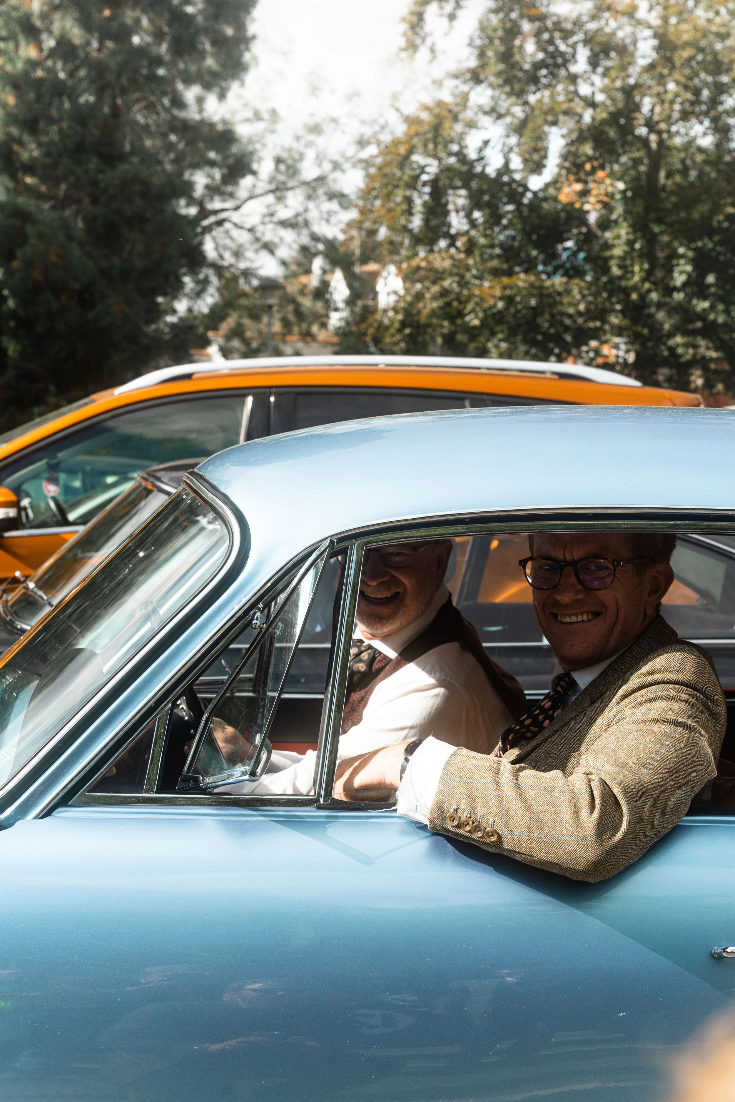 Two men dressed in vintage clothing sitting in a classic blue car, smiling and looking towards the camera, with another vintage orange car behind them, outdoors with trees and a sunny sky in the background.