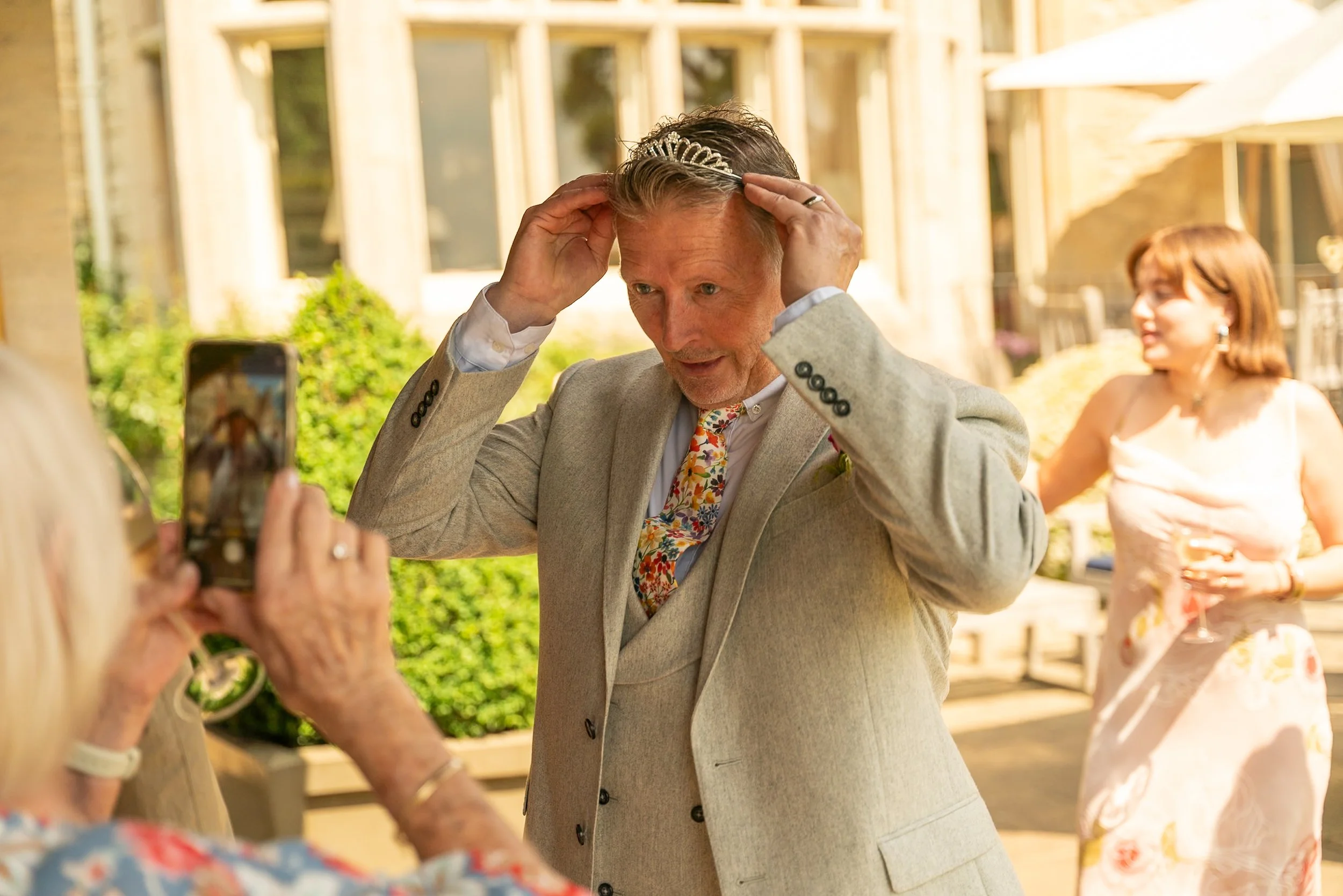 Man adjusting a tiara on his head at a social gathering outdoors, with another woman taking a photo of him, and a woman holding a drink in the background.