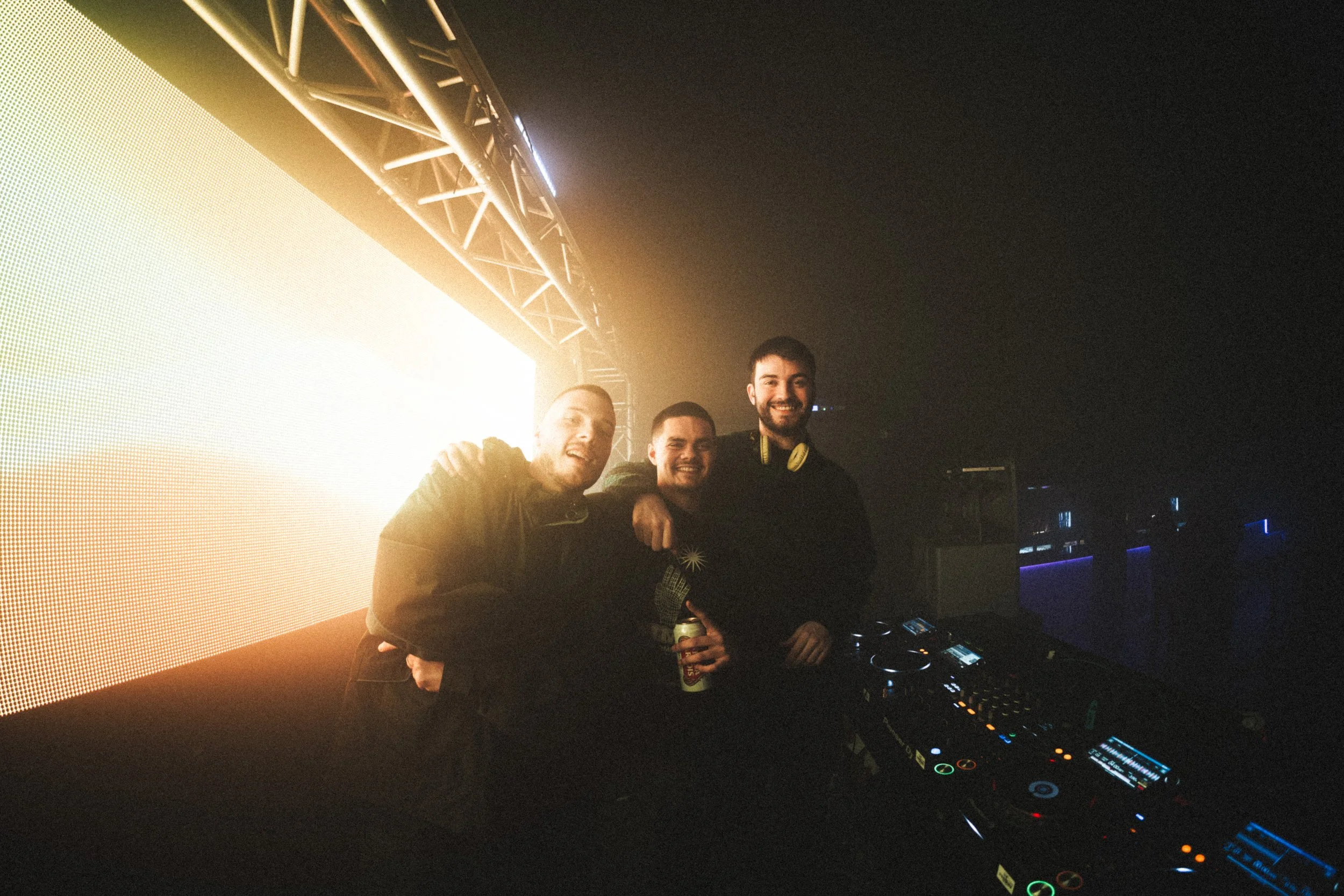 Three young men in a DJ booth at a nightclub or music event, smiling and posing for the camera with DJ equipment in front of them. The lighting is dark with bright stage lights in the background.
