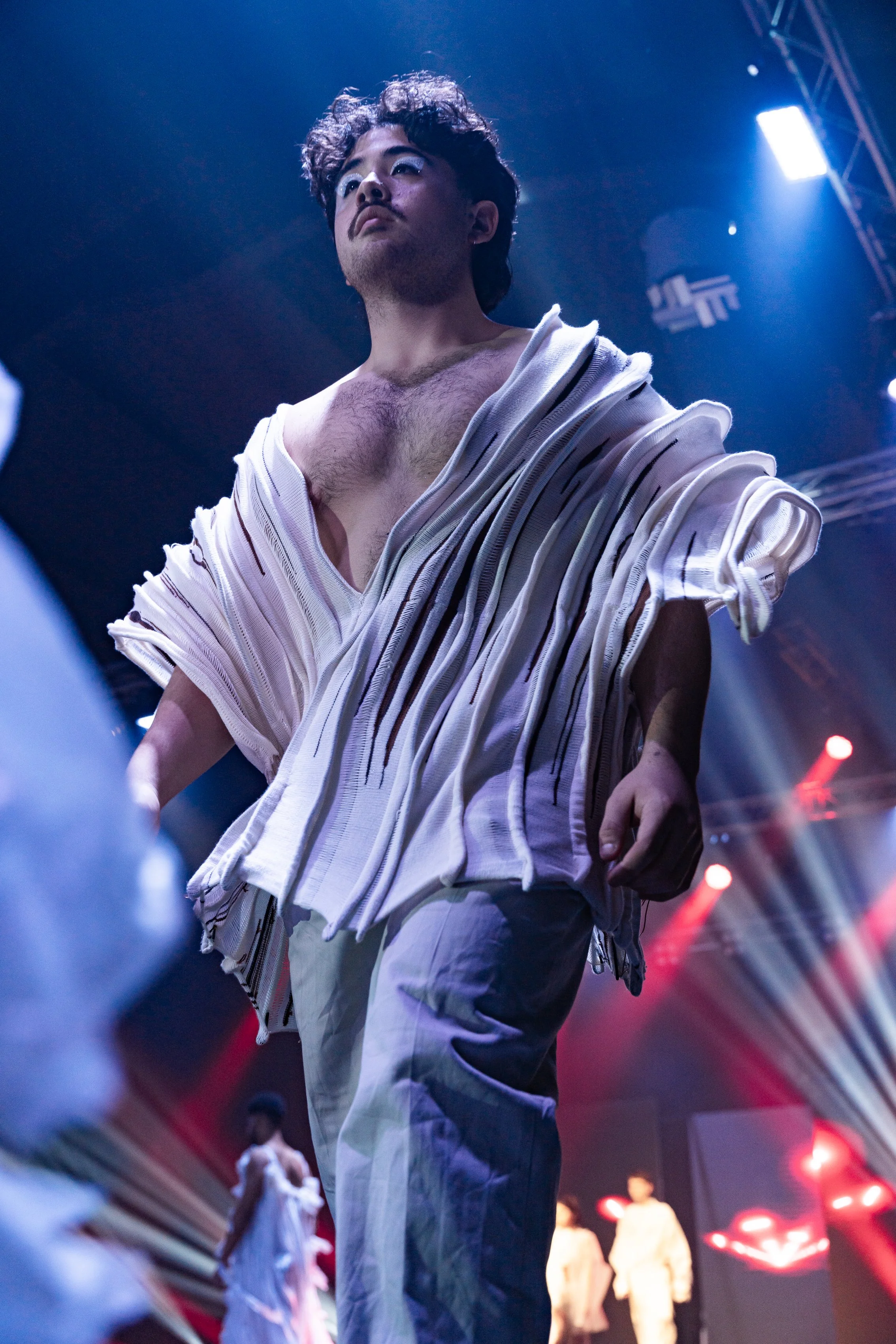 A male model walking on a runway during a fashion show, wearing a unique white garment with layered, striped fabric, with bright stage lights overhead.