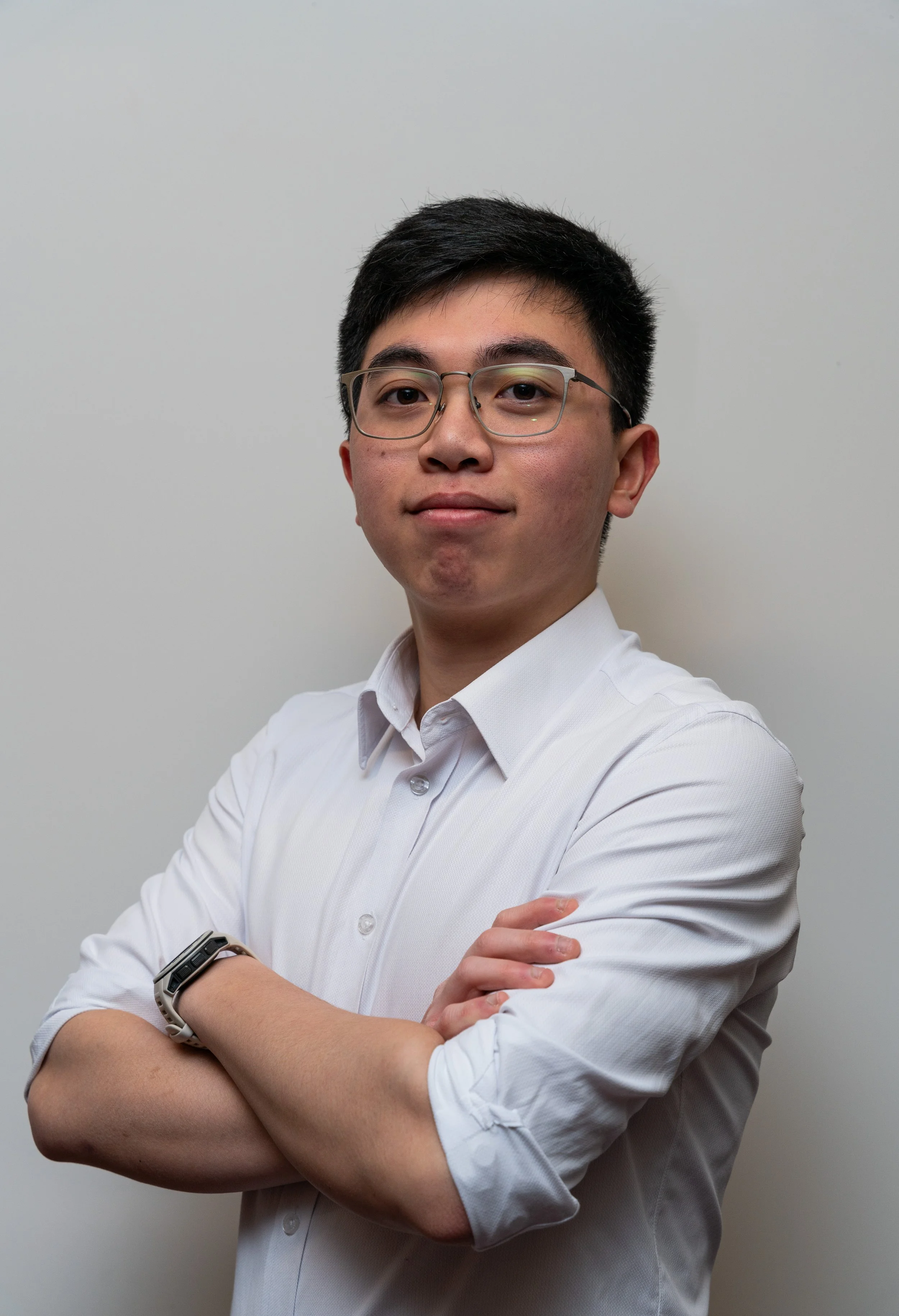 Young man with glasses, dressed in a white shirt, standing against a plain wall, arms crossed, looking at the camera.