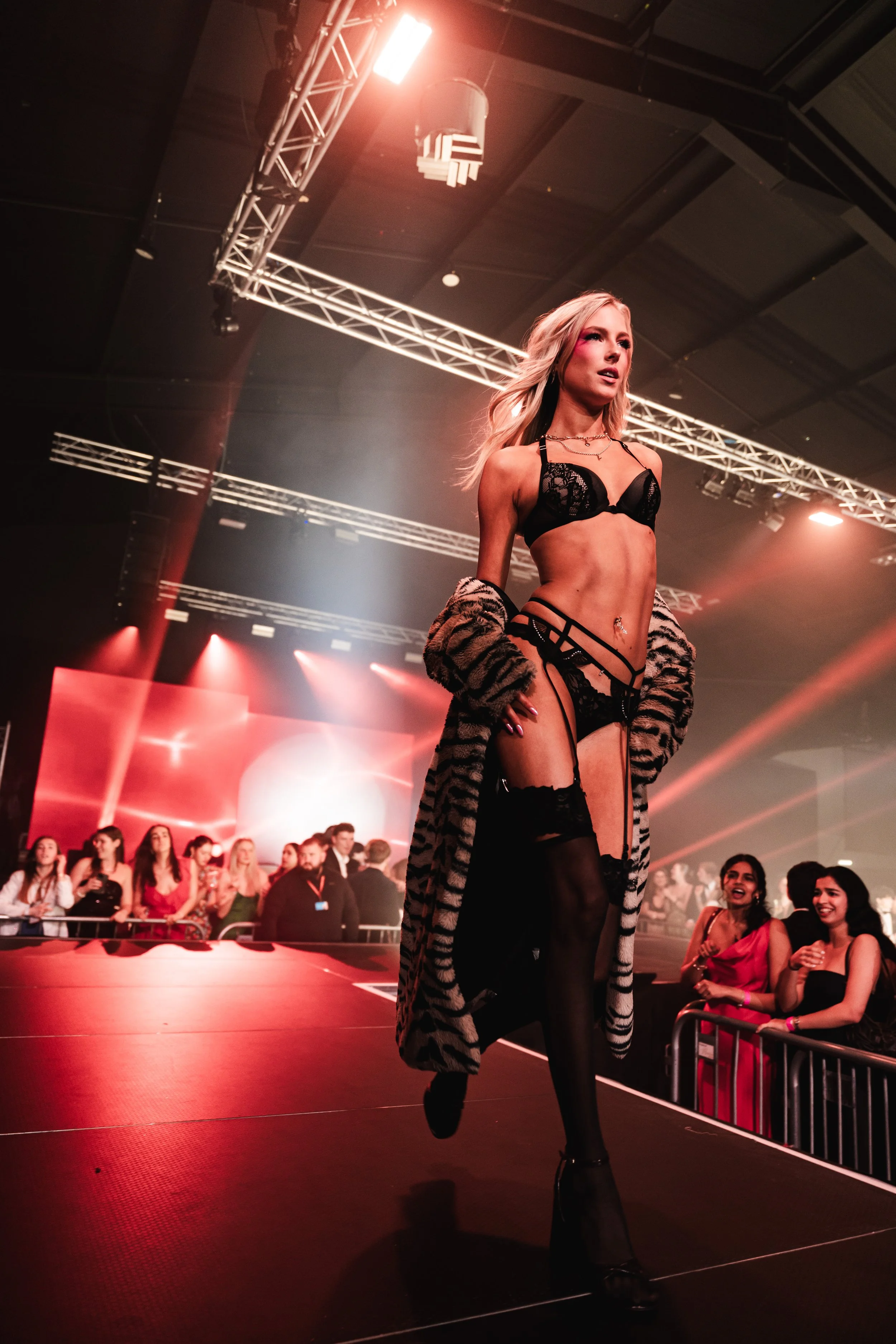 A woman walking on a runway at a fashion show, wearing black lingerie, a striped coat, and thigh-high stockings, with an audience in the background.