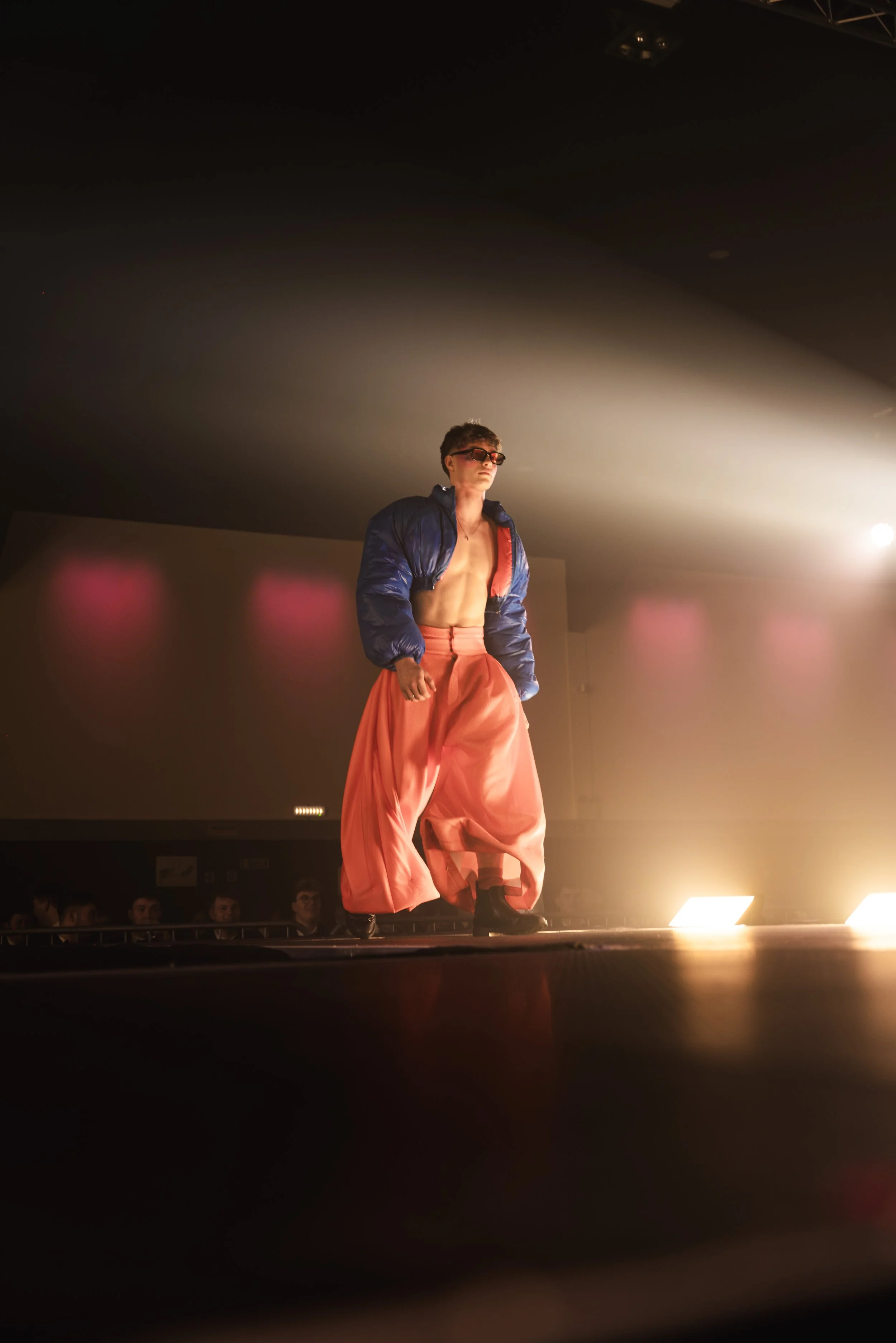 Male model walking on runway wearing large pink baggy pants, a dark blue puffer jacket, sunglasses, and black shoes in a fashion show setting with dramatic lighting.