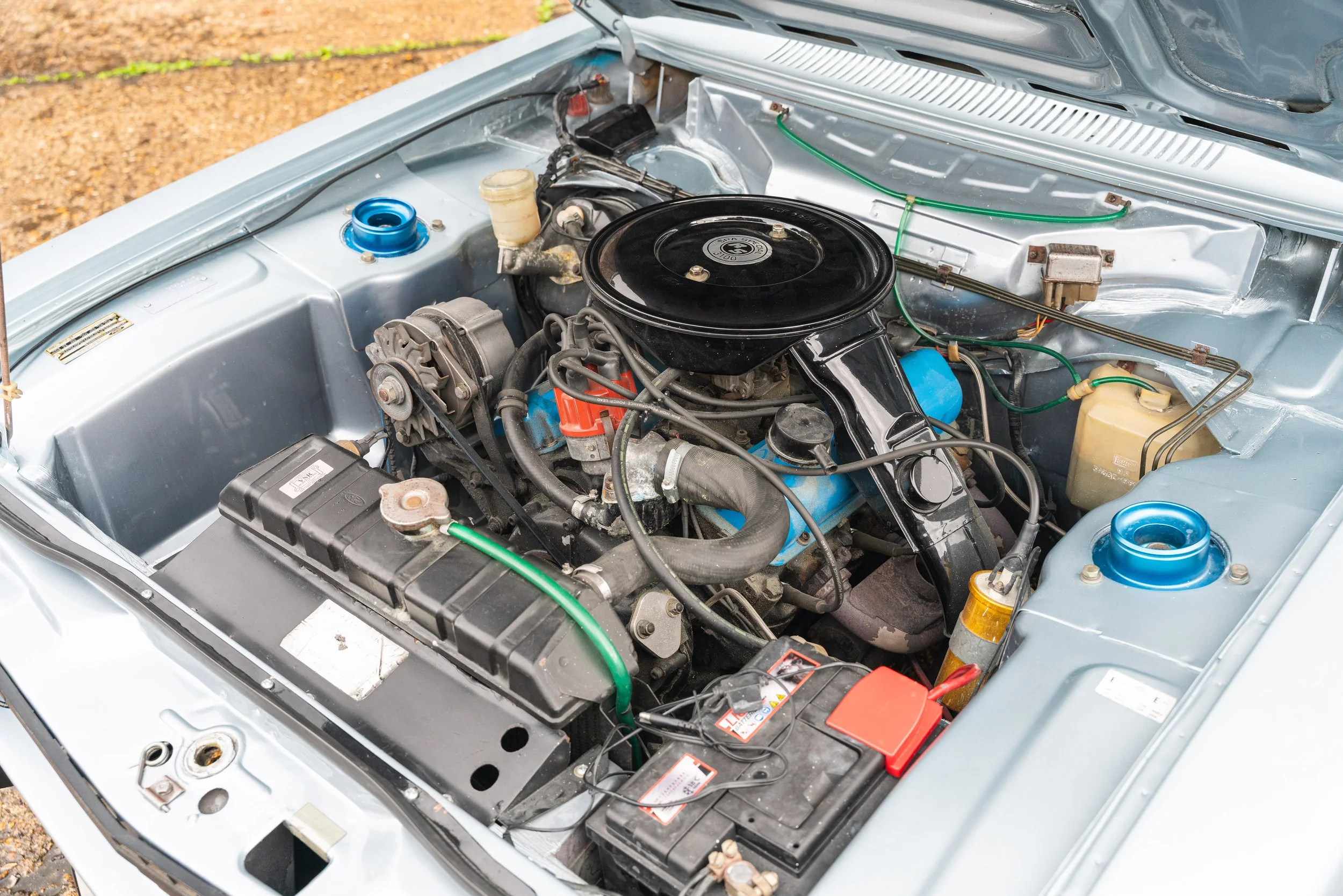 Engine bay of a vintage car with various components including a black round air filter, hoses, and a battery.