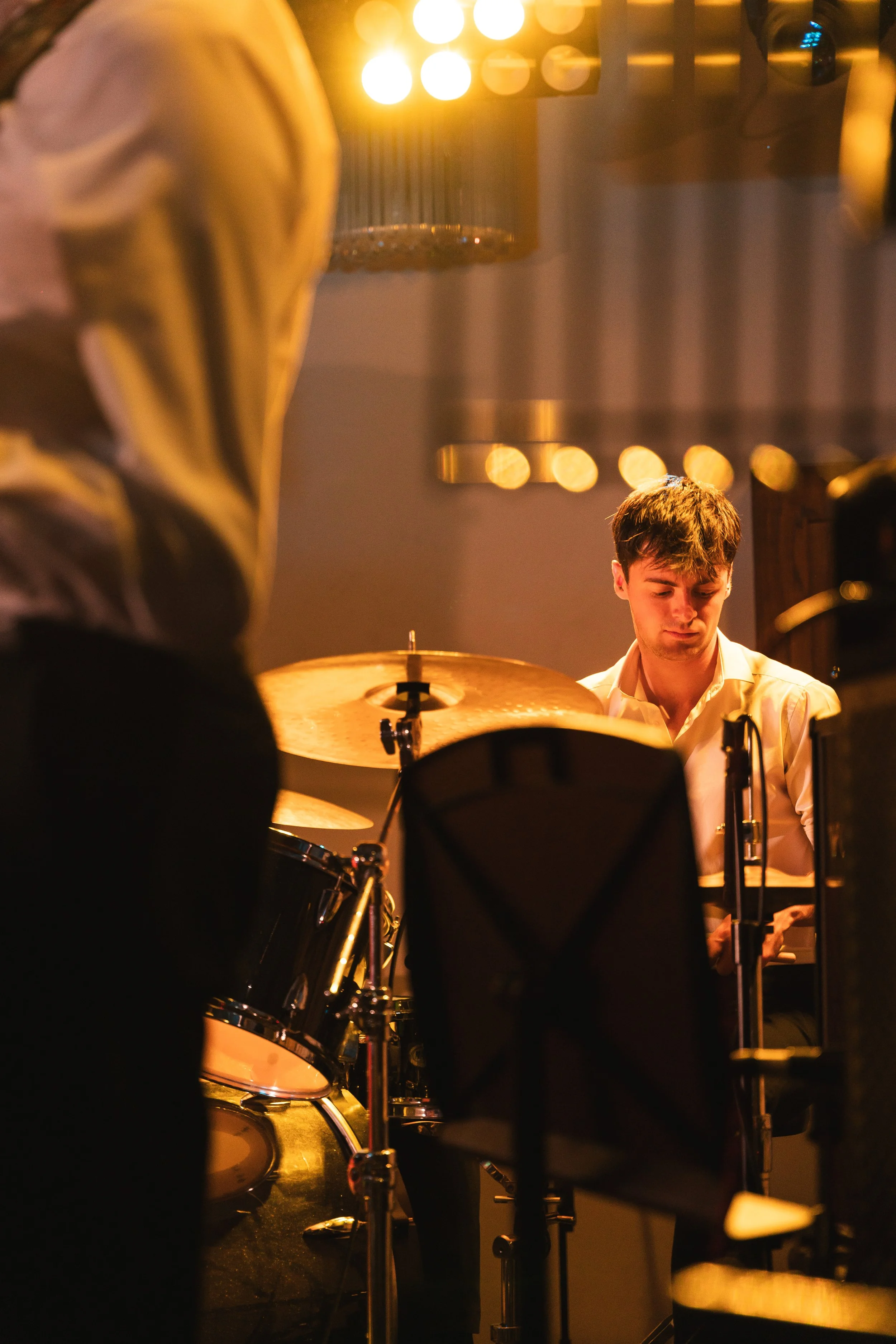 A young man playing the drums during a live performance in a dimly lit venue with warm lighting and blurred circular light reflections in the background.