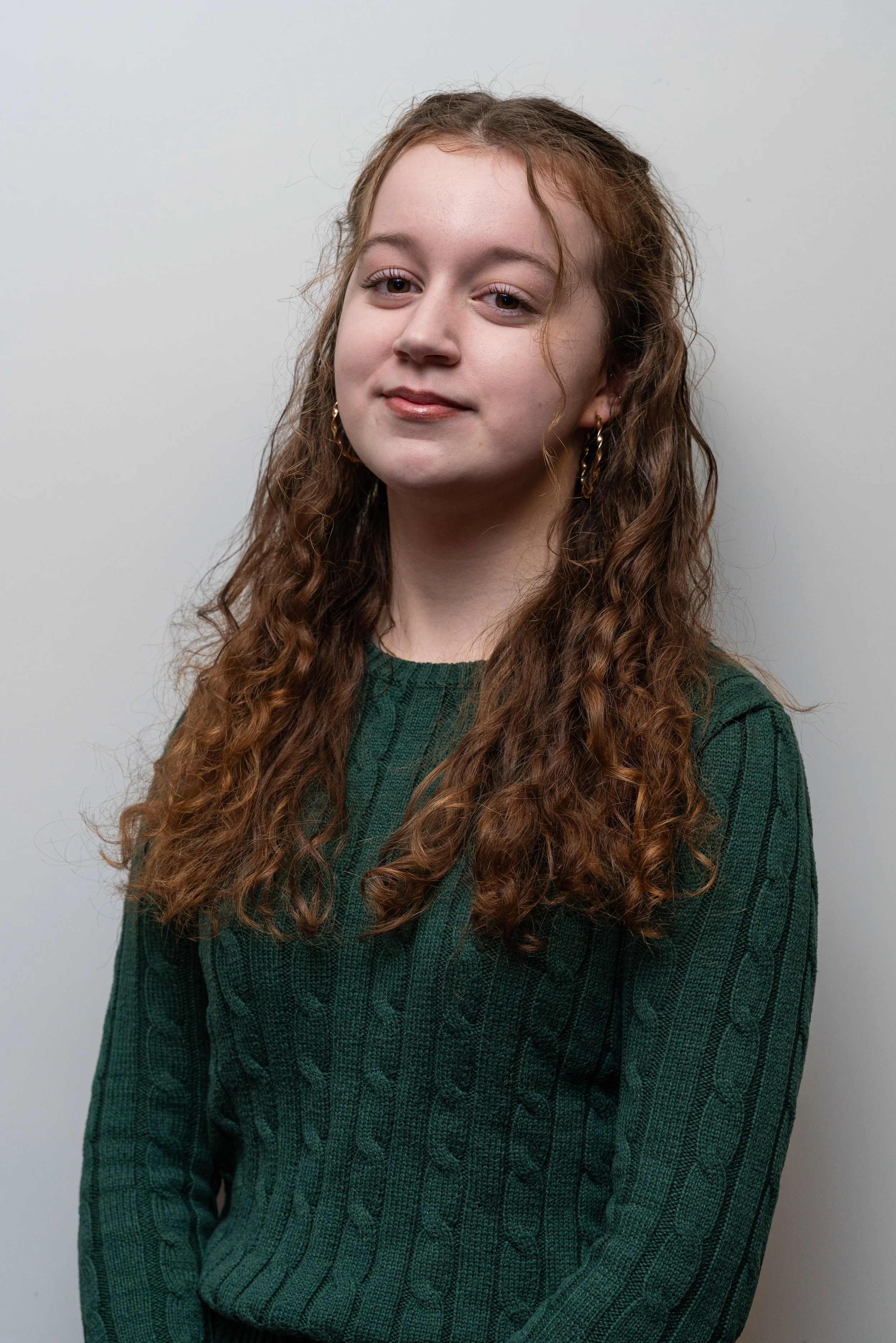 A young woman with long, curly red hair and hoop earrings, wearing a dark green cable-knit sweater, standing against a light gray background.