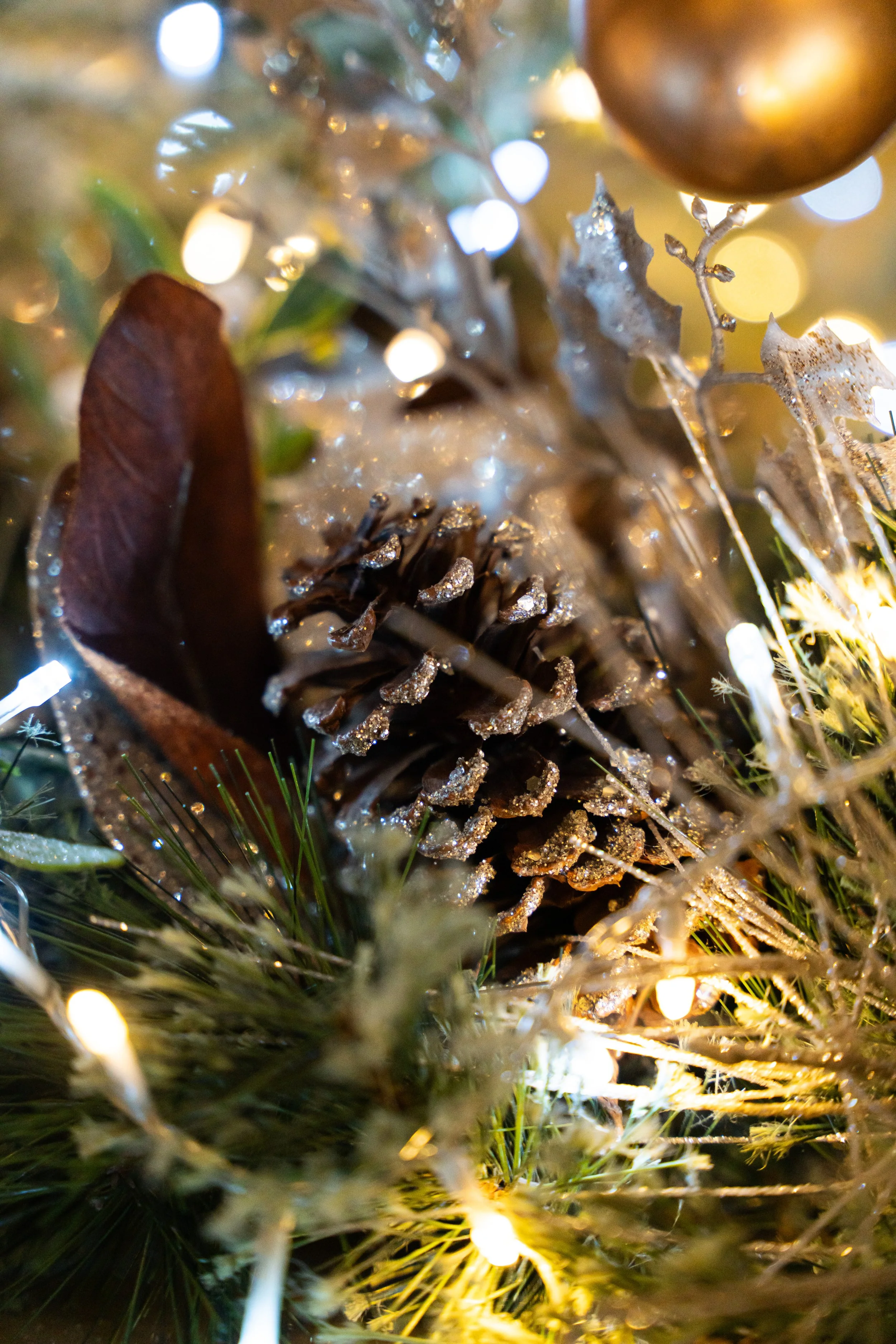 Close-up of a decorated Christmas tree branch with pinecones, glittery leaves, and warm white string lights.