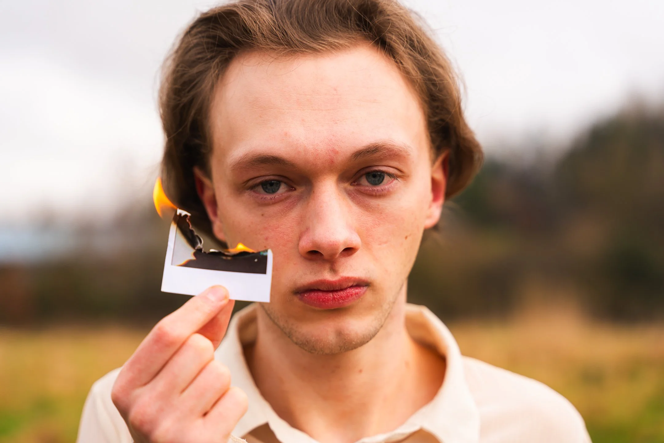 A young man with light skin, light brown hair, and blue eyes holding a burnt photo print with a small flame near the edge, outdoors in a blurred natural setting.