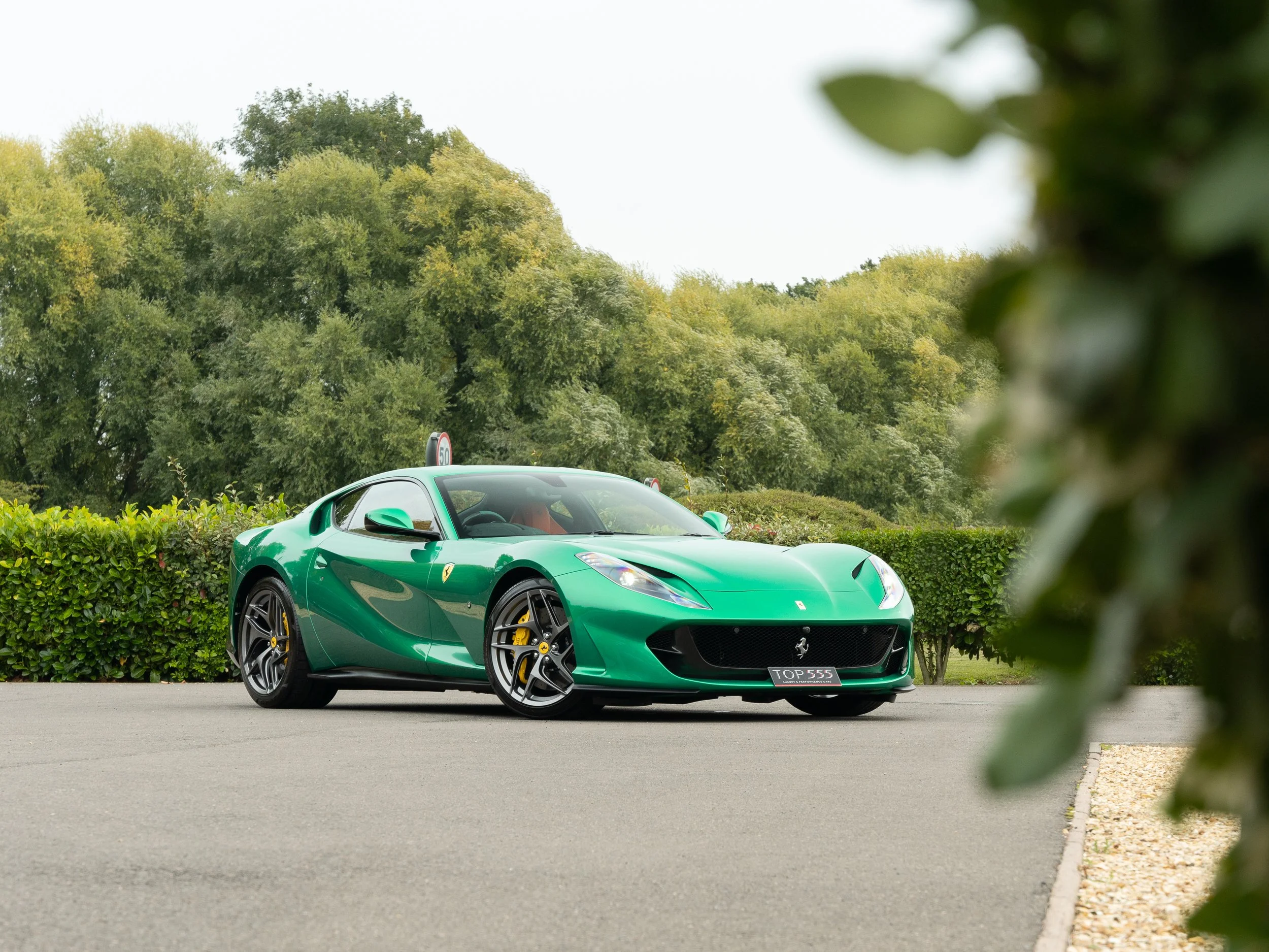 A green Ferrari sports car parked on a paved surface with trees and bushes in the background.