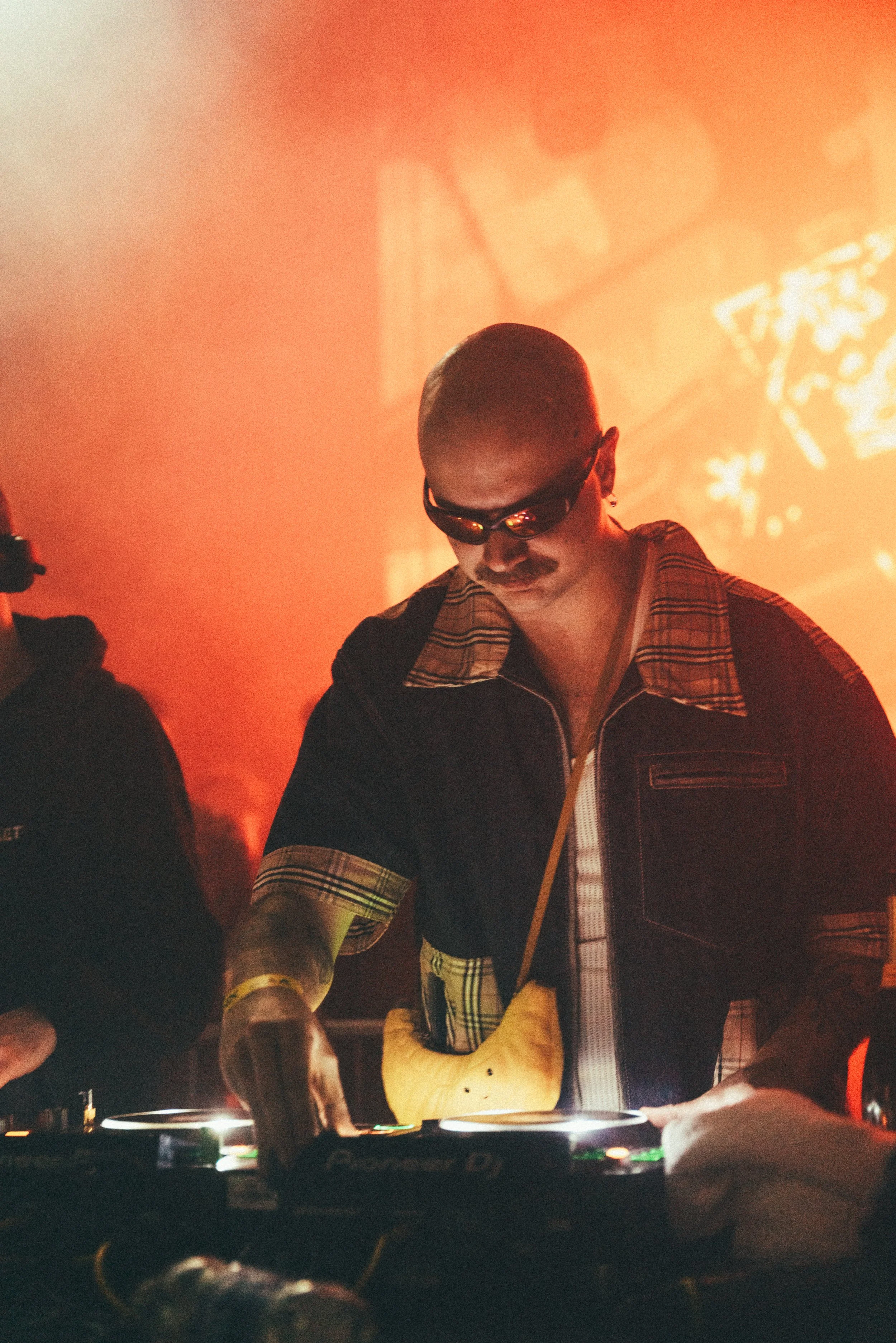 A DJ with sunglasses and a plaid shirt DJing at a nightclub with warm orange lighting and shadows in the background.