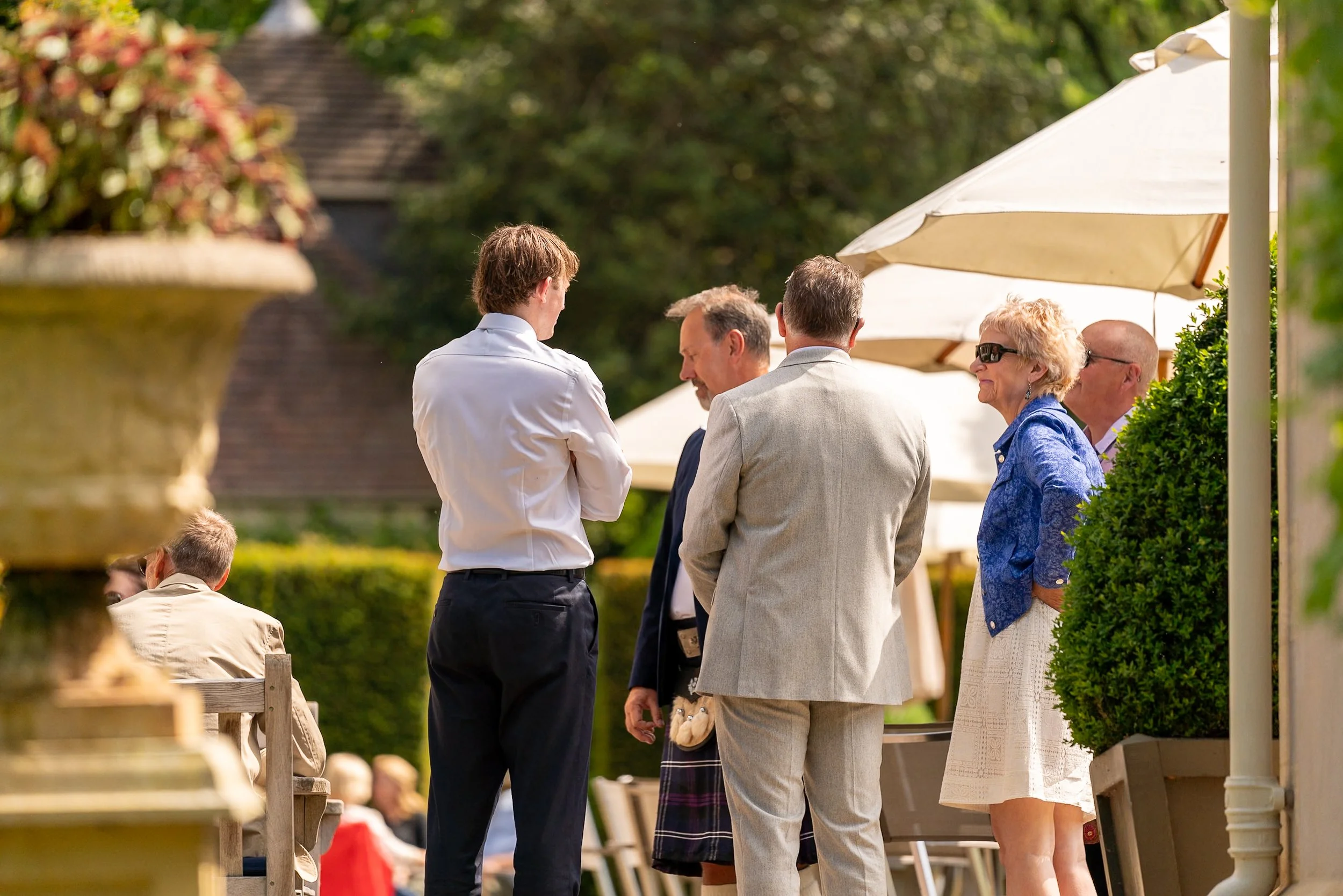 Group of six people standing outdoors, talking and enjoying sunny weather, with some wearing formal or semi-formal clothing, under umbrellas.