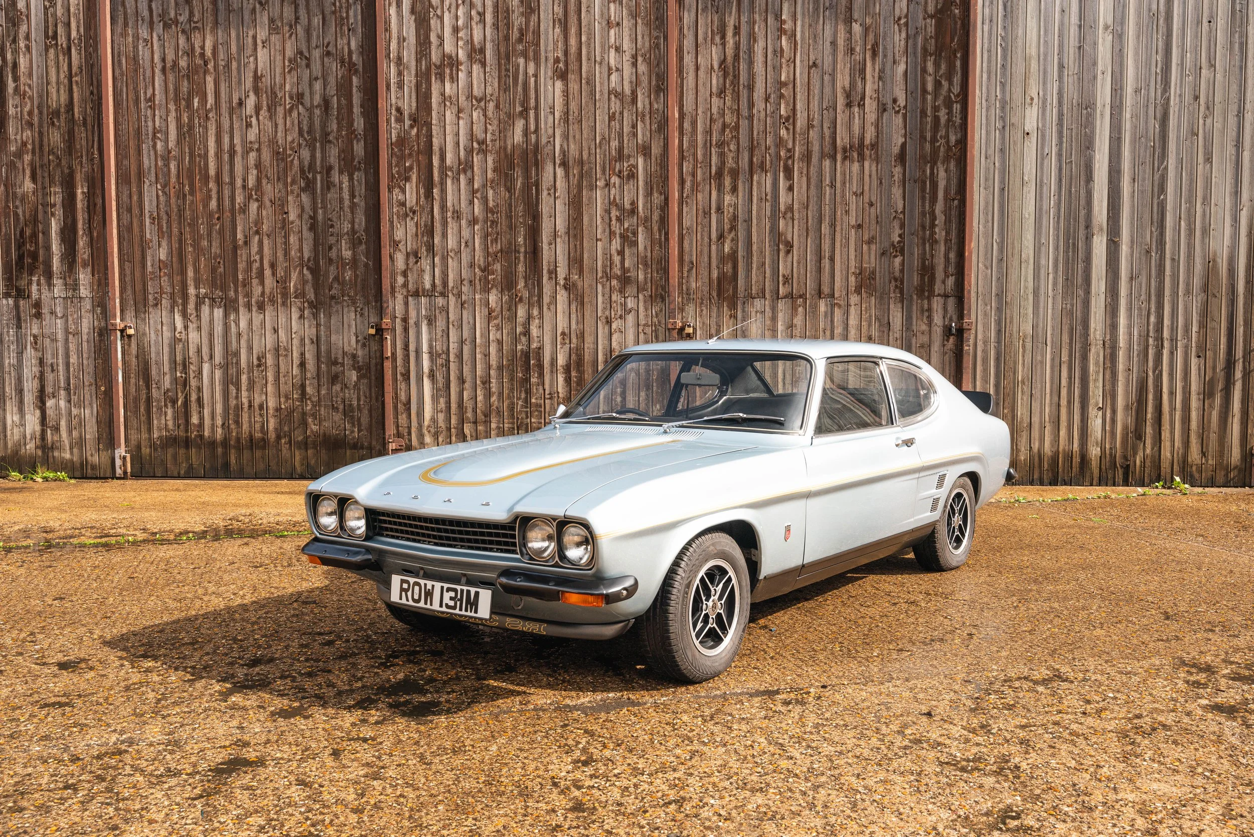 A vintage silver sports car with a black grille and wheels, parked on gravel in front of a large wooden barn door.