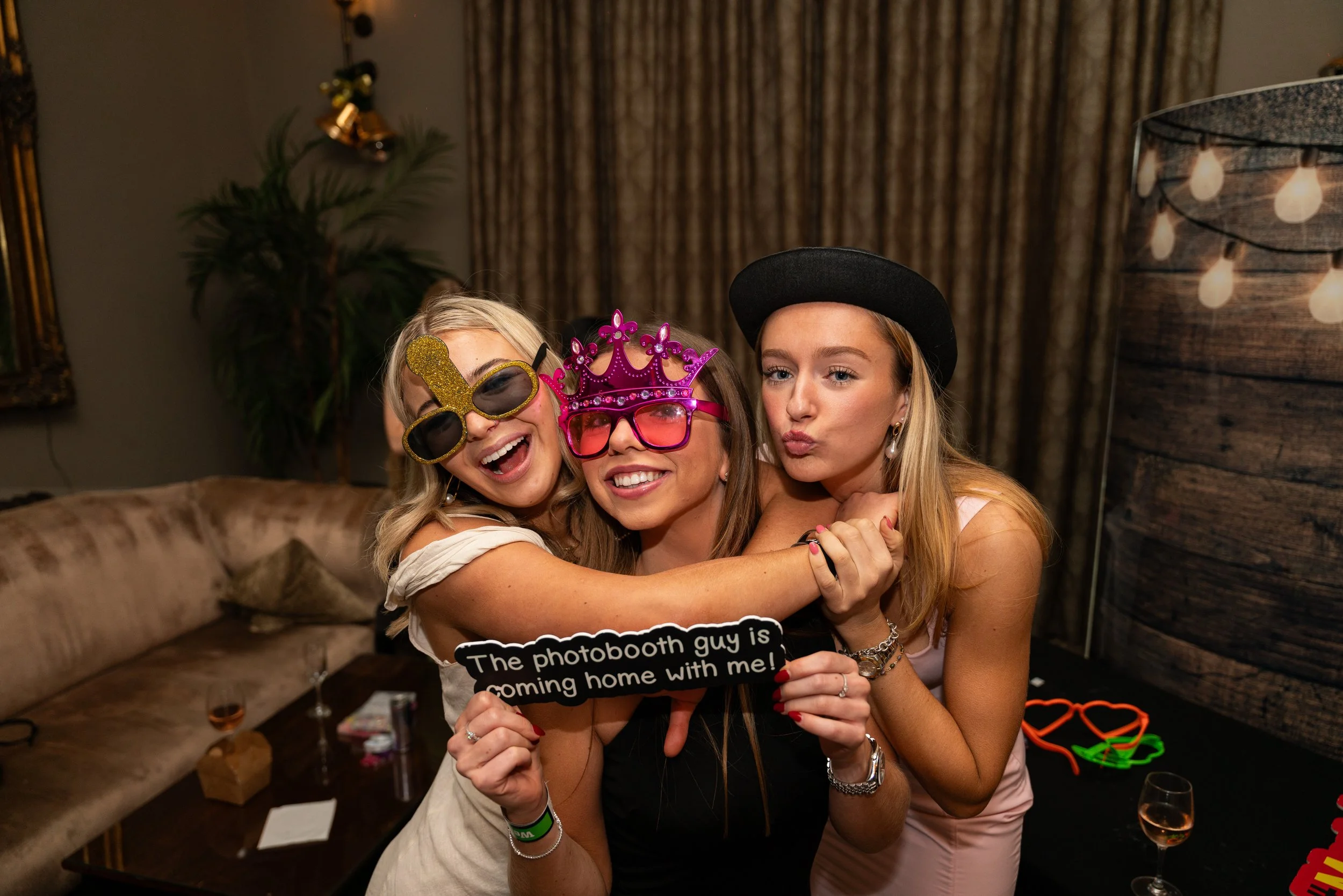 Three women celebrating at a party wearing funny glasses and holding a sign that says "The photobooth guy is coming home with me!" in a living room decorated for a celebration.
