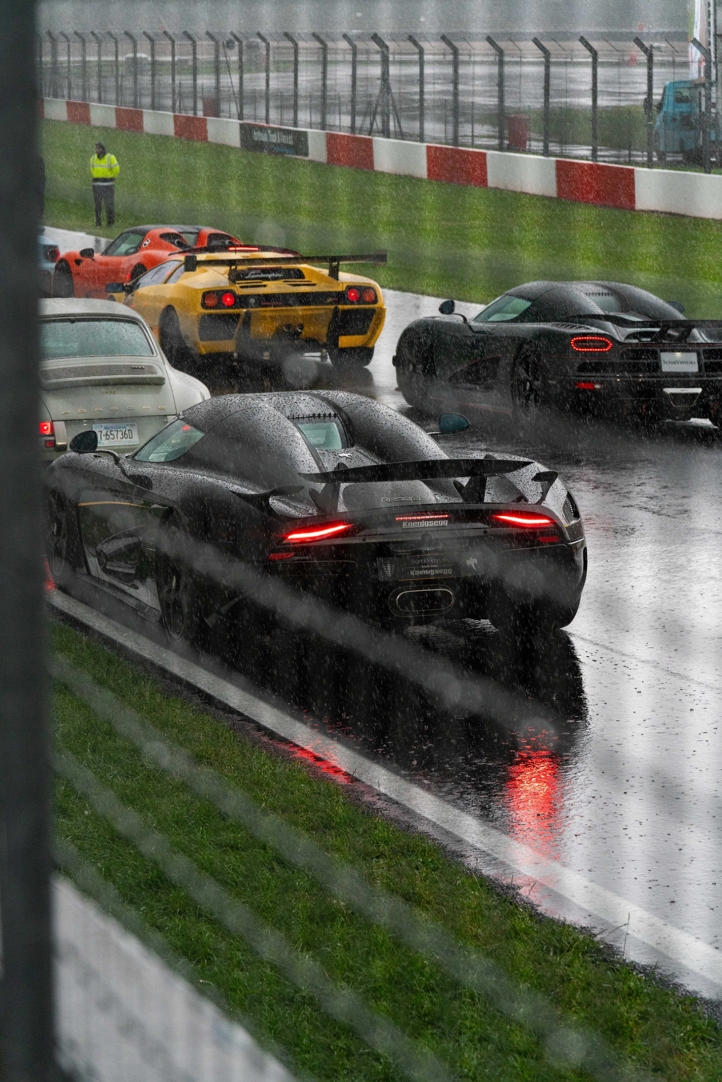 Multiple sports cars parked on a wet racetrack during rainy weather, with a grassy barrier and a person standing in the background.