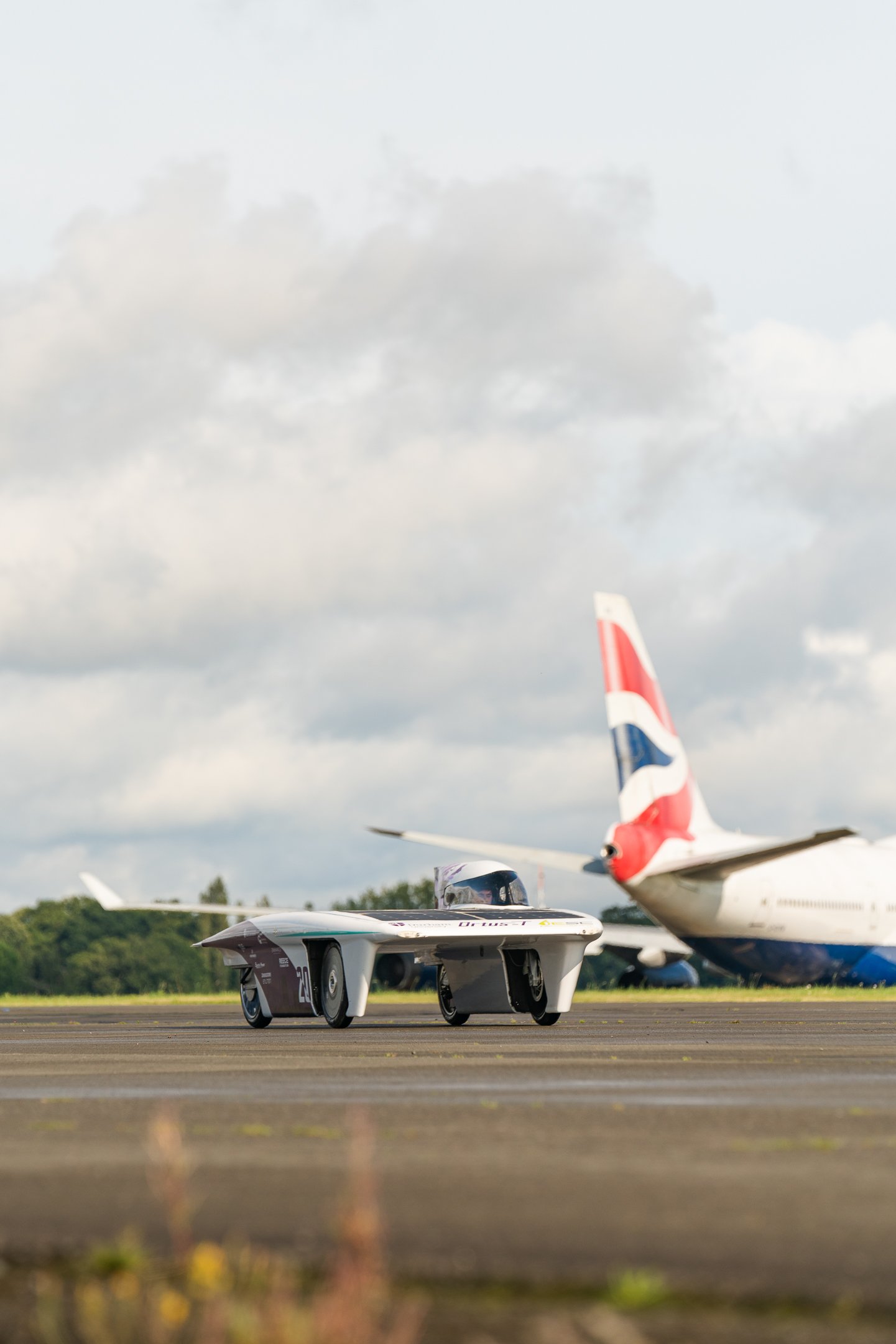 A solar-powered race car on airport tarmac with British Airways airplane in the background.