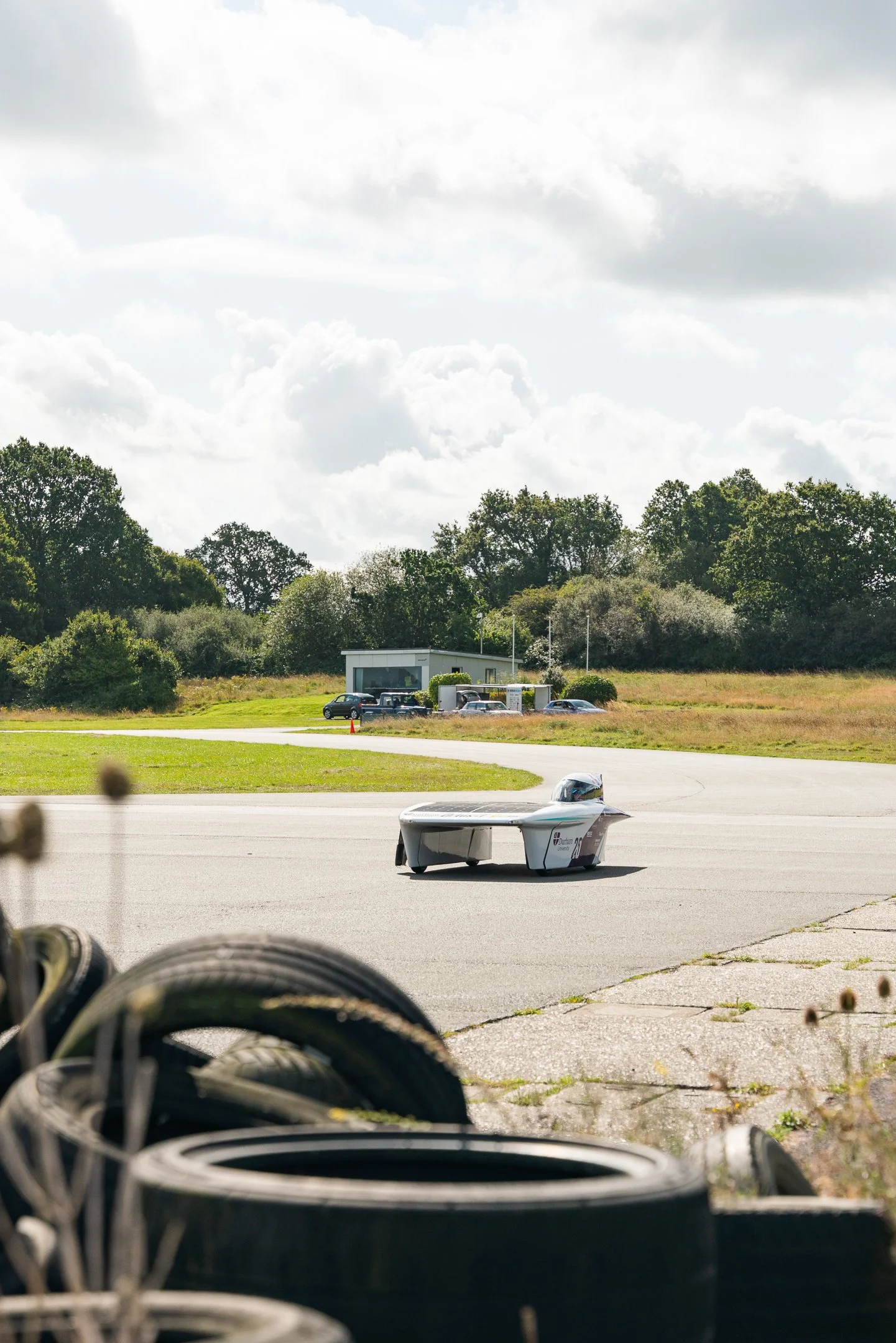 A solar-powered race car on a tarmac at an outdoor track with a building, parked cars, and greenery in the background under a partly cloudy sky.