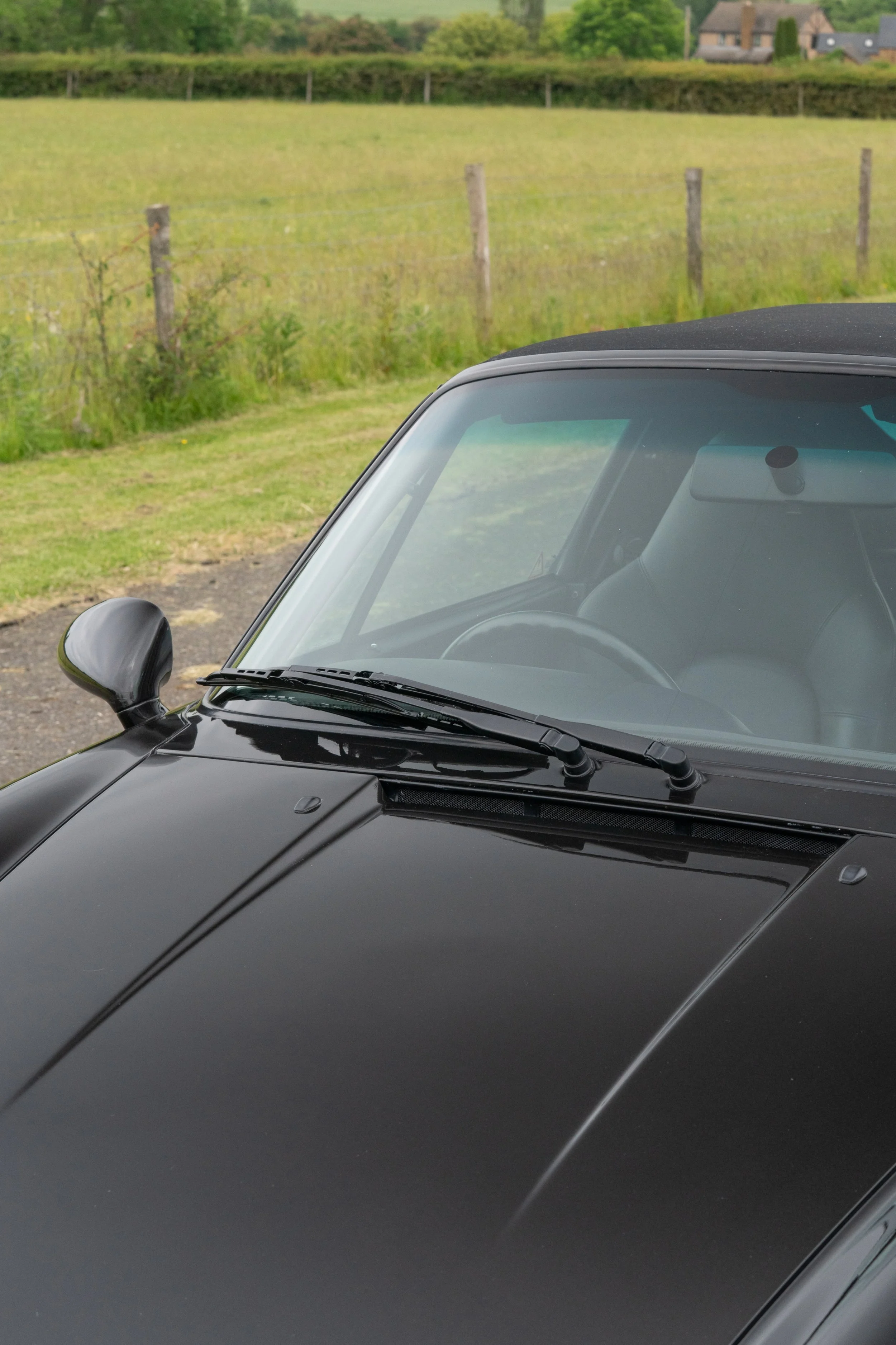 Close-up of a black sports car's windshield, side mirror, and hood with a scenic rural background.