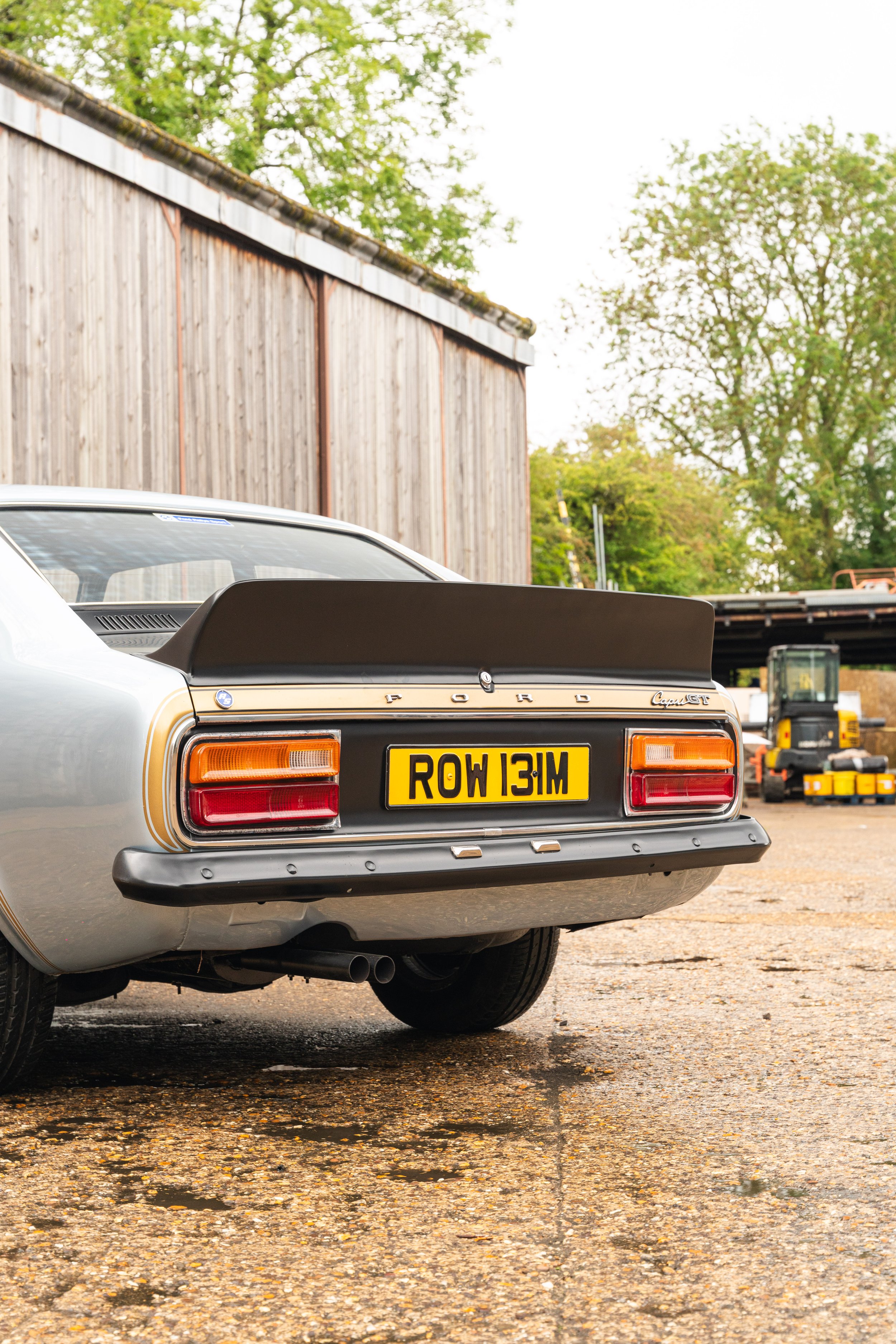 Rear view of a vintage Ford Capri car with a black spoiler and a yellow license plate reading 'ROW 131M,' parked on a gravel surface near a wooden building with trees in the background.