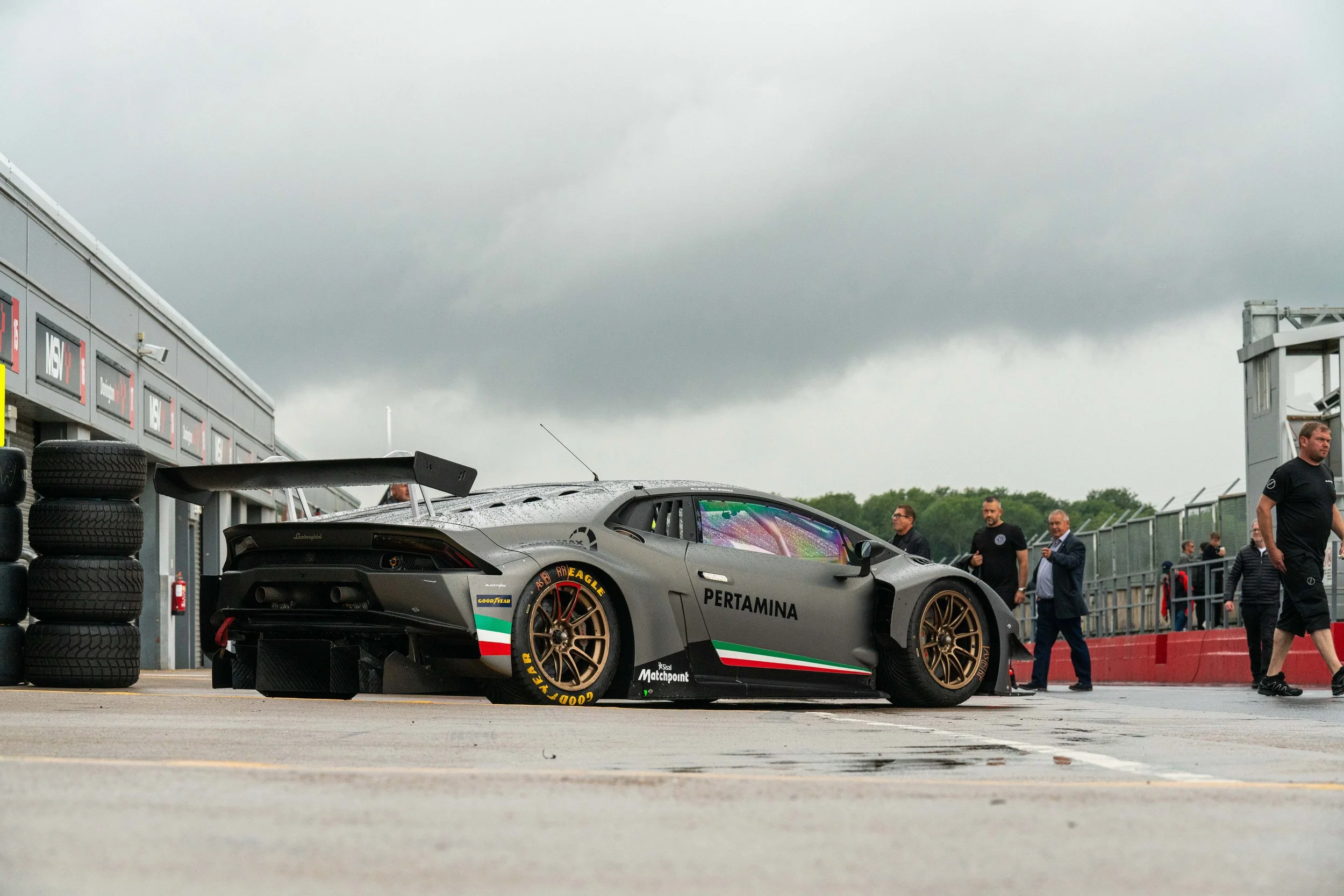 A sleek, matte gray Lamborghini race car in the pit lane of a racetrack, with a large rear wing, gold wheels, and sponsor logos including 'PERTAMINA', under a cloudy sky, surrounded by team personnel and officials.
