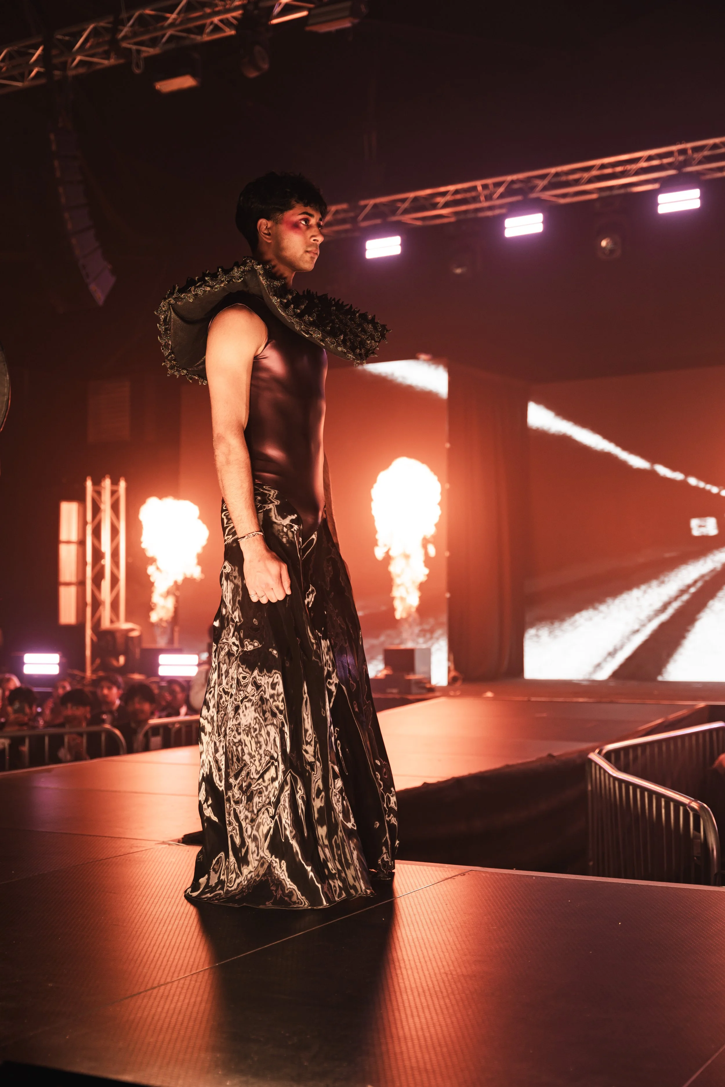 A male model walks on a fashion runway with dramatic lighting, wearing a black sleeveless top and patterned wide-leg pants, with fiery stage lights in the background.
