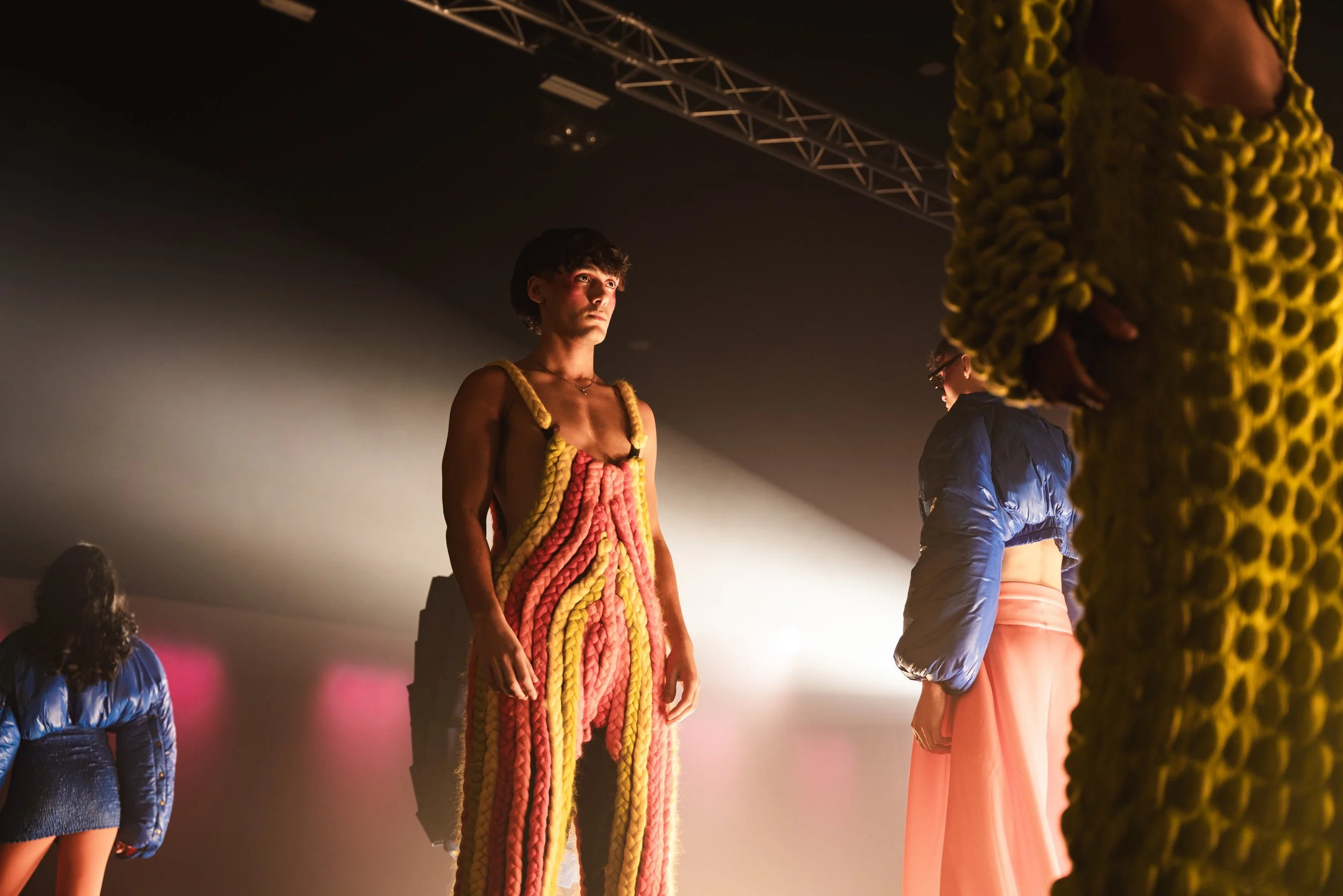 Models on runway wearing colorful, textured clothing in a dimly lit setting.