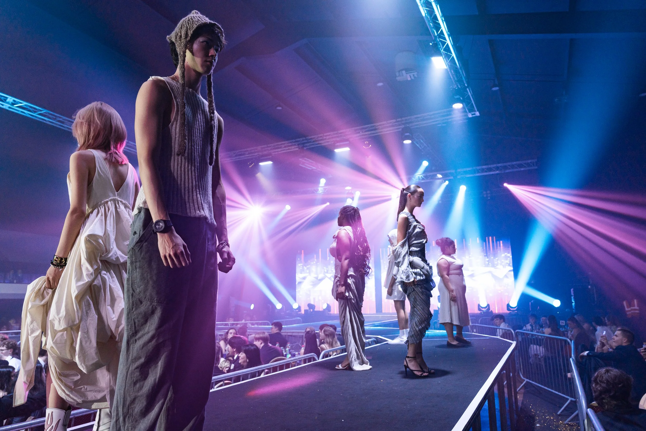 Multiple models standing on a runway during a fashion show, with colorful stage lighting and an audience watching.