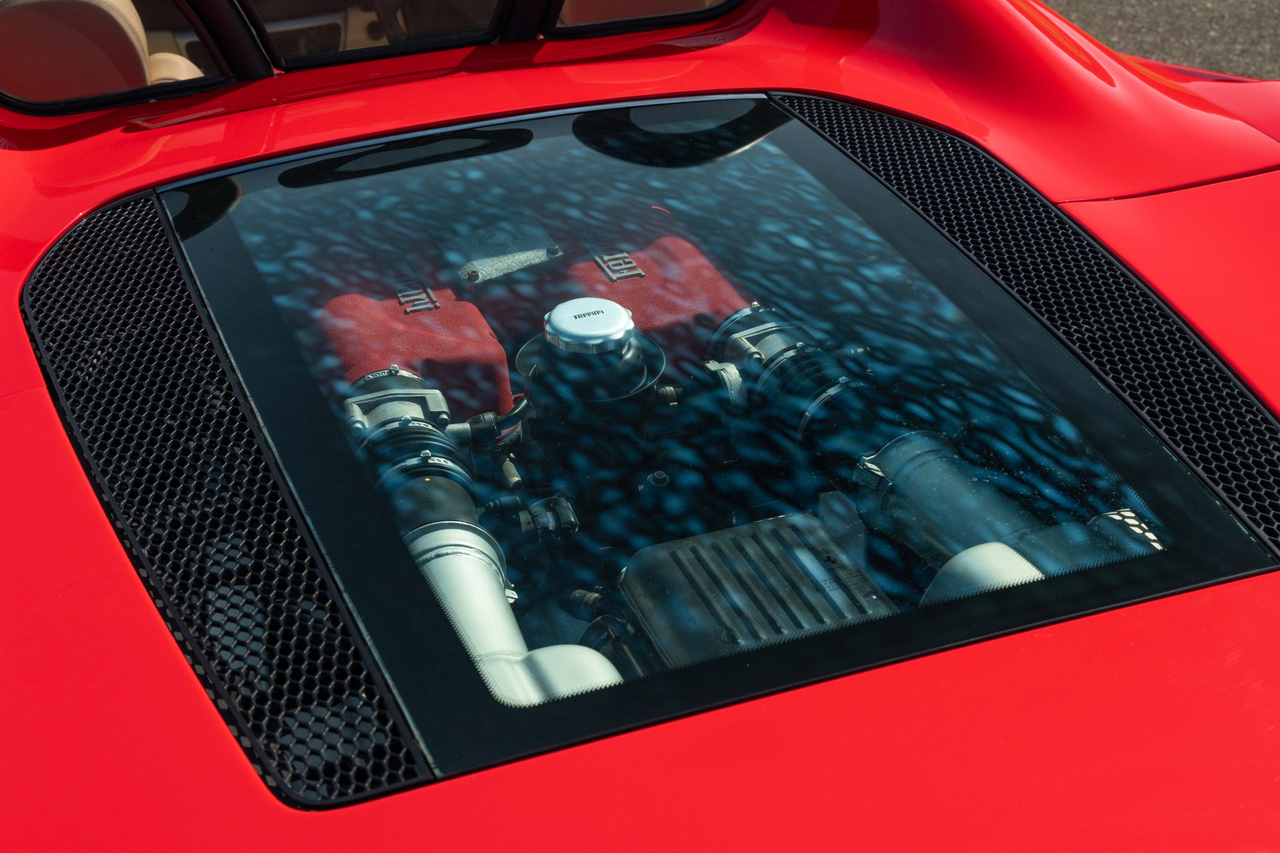 Close-up of a red sports car's engine visible through a clear rear window, showing Ferrari branded engine components.