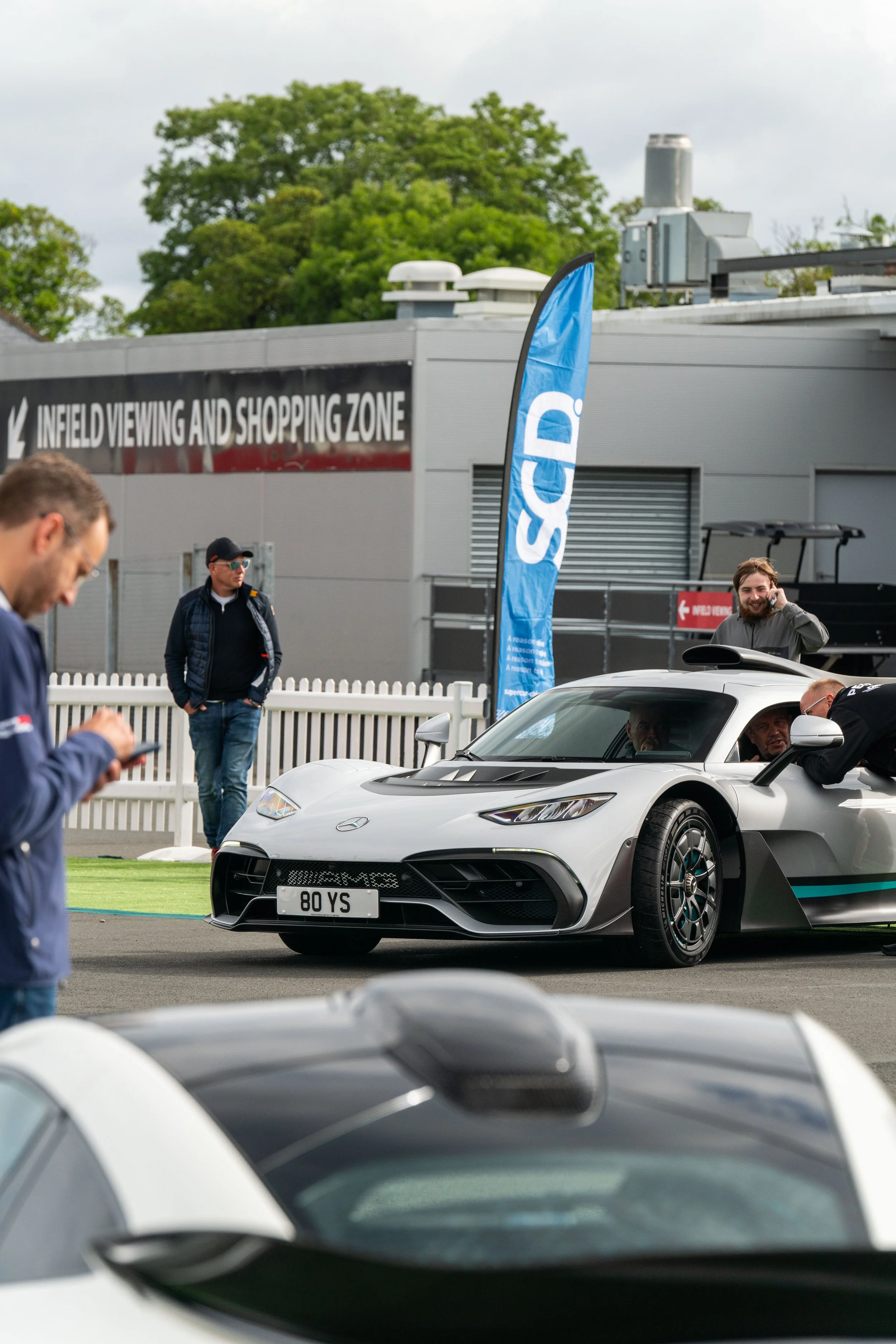 A silver sports car with a license plate reading '80 YS' parked near a group of people at a racing event, with a blue flag and a sign in the background.