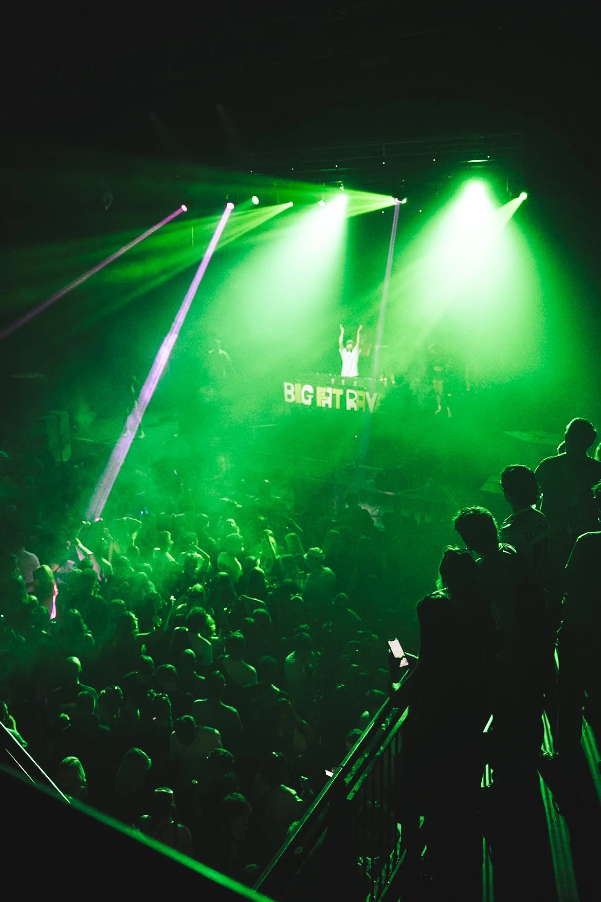 A crowded dance floor at a concert with green stage lighting and a DJ on a raised platform.