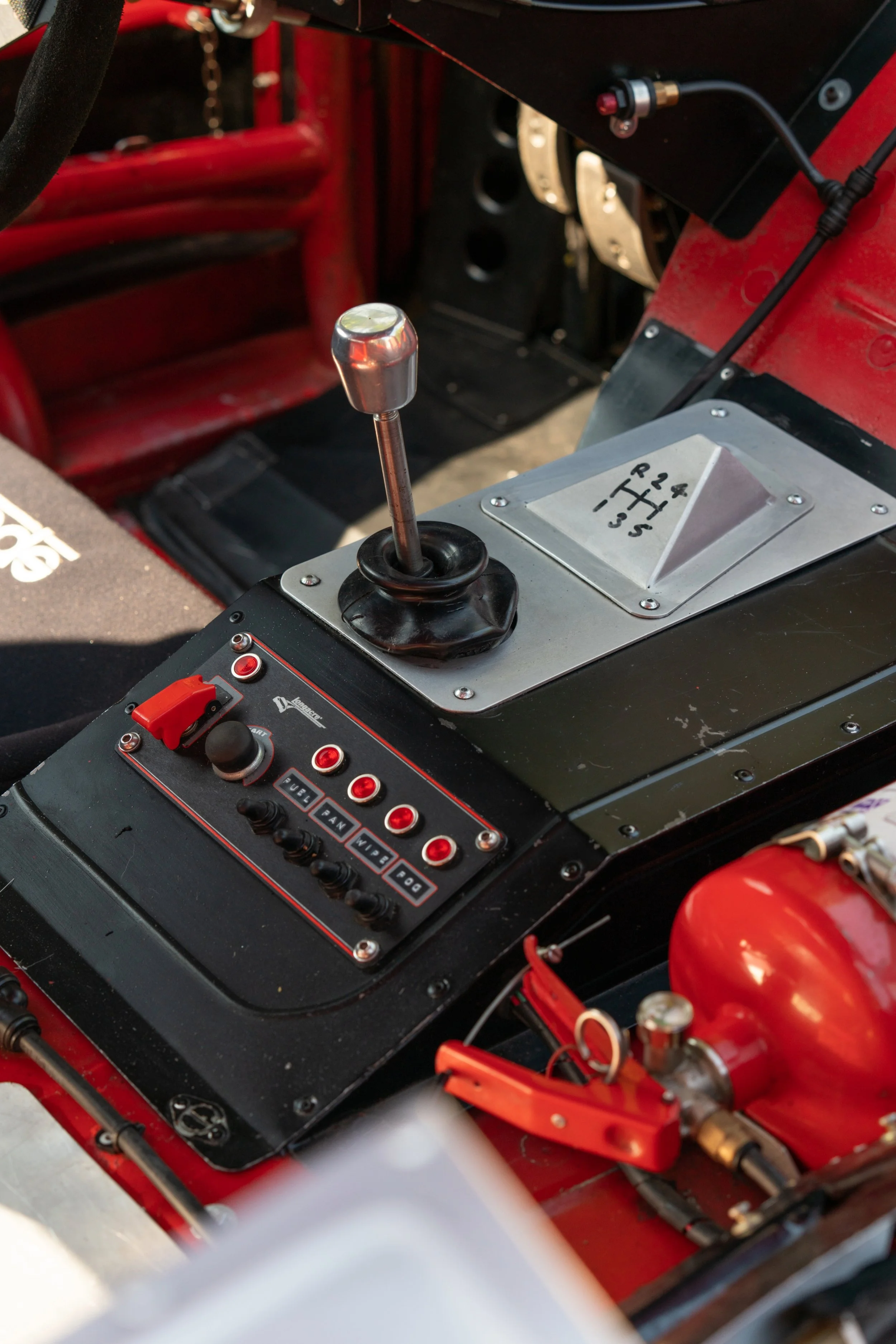 Close-up of the gear shift and control panel inside a race car, featuring a manual gear shifter, red fire extinguisher, and various switches and buttons.