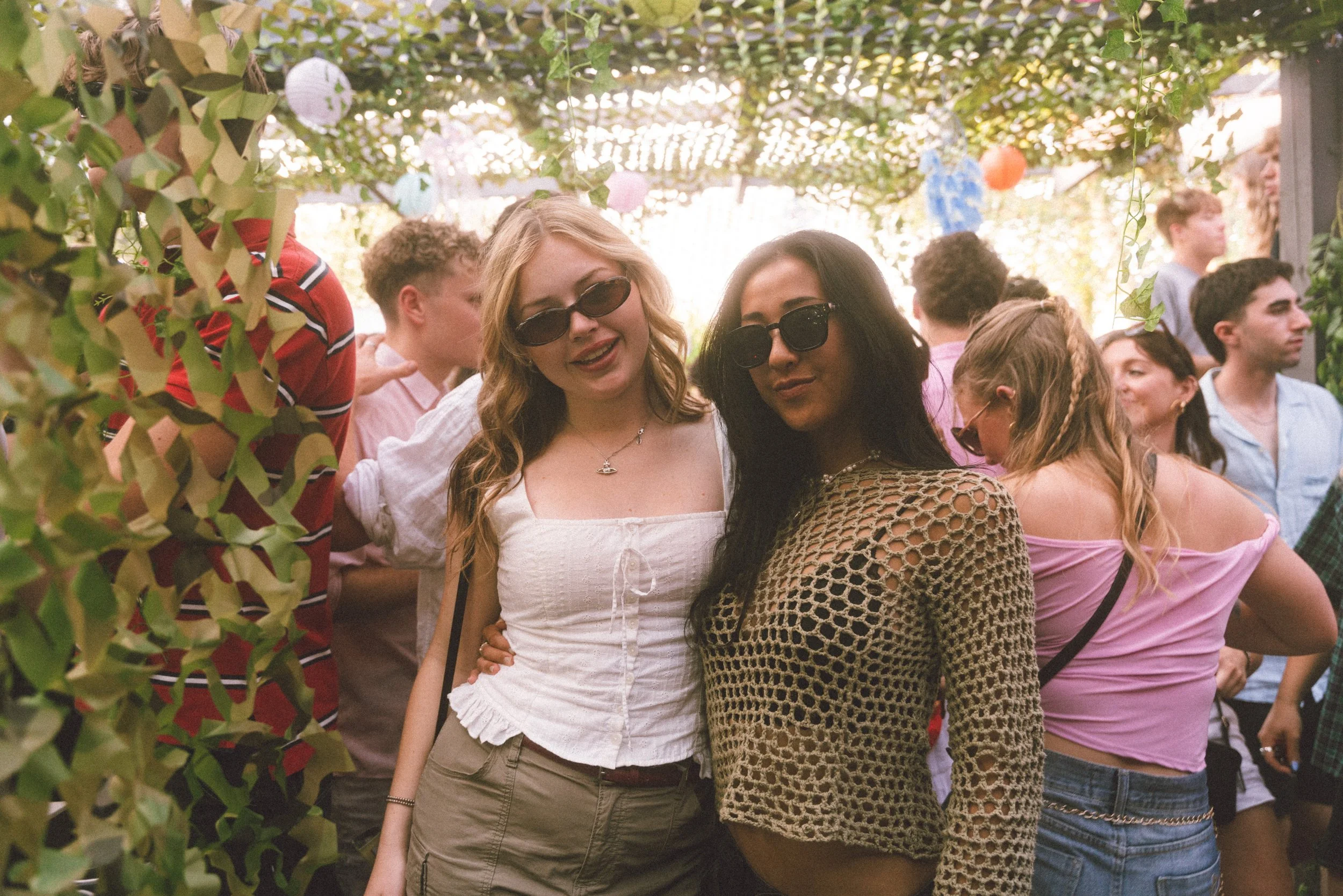 Two women wearing sunglasses and fashionable outfits smiling at the camera at a social gathering or party, with other people in the background.