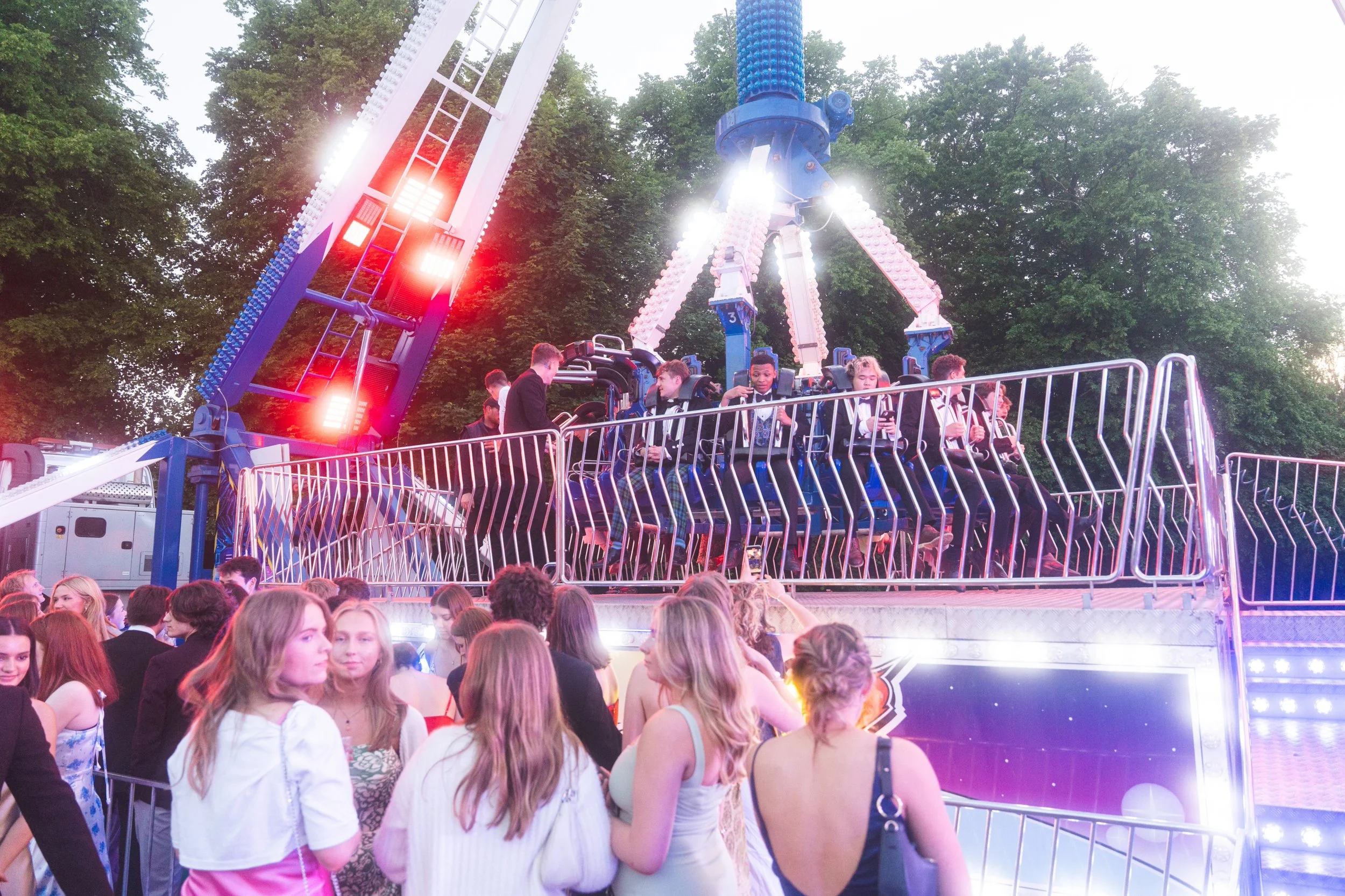 A lively scene at a fair with people waiting in line for a blue amusement park ride, with some riders seated and others standing nearby, and a green, tree-filled park in the background.