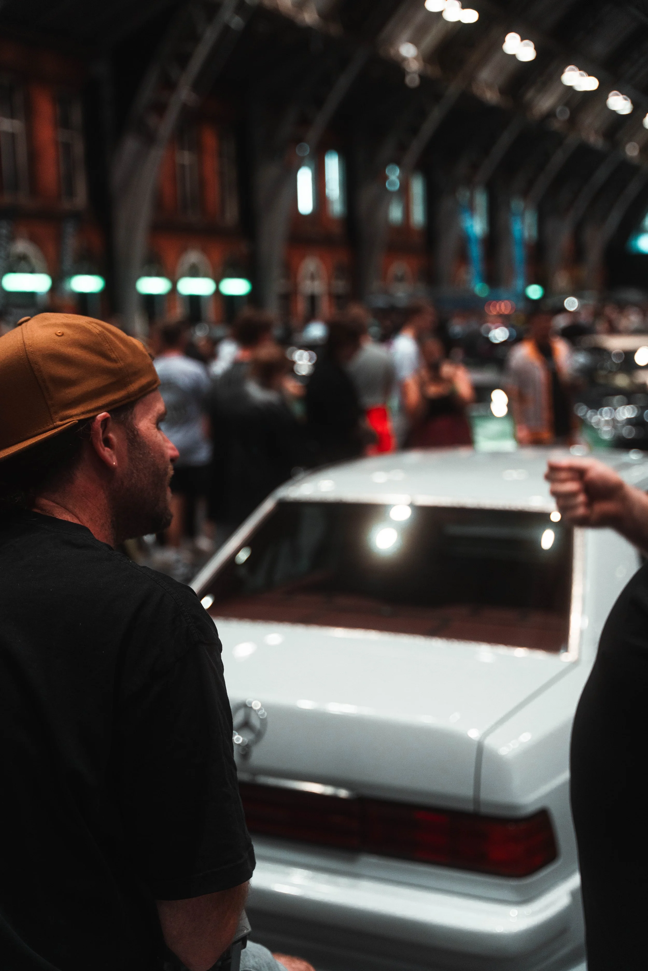 A man with a tan cap sits next to a white classic Mercedes-Benz in a crowded indoor venue with arched ceiling and people in the background.