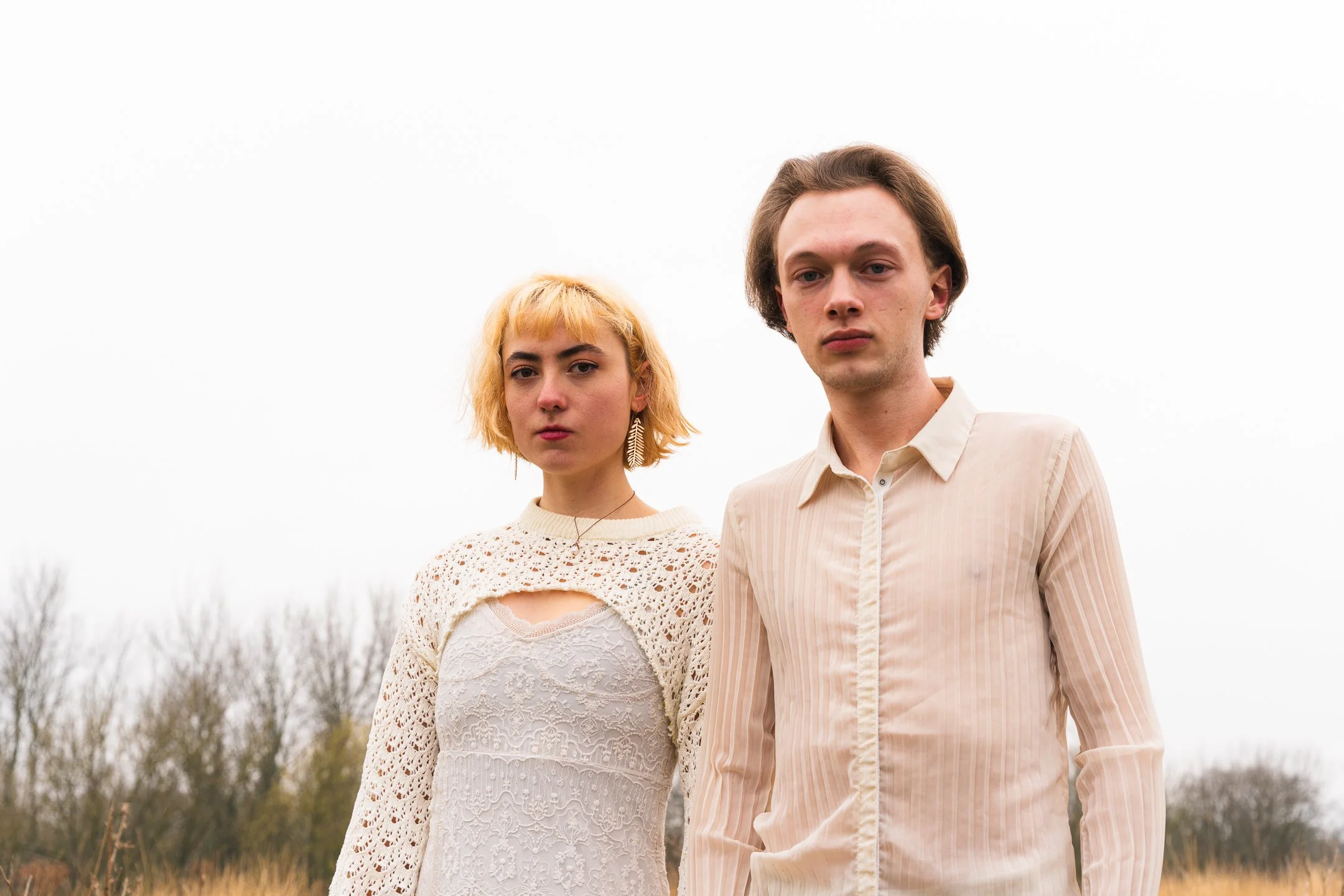 A young woman with short blonde hair and a young man with medium-length brown hair standing outdoors on a cloudy day, both looking serious.