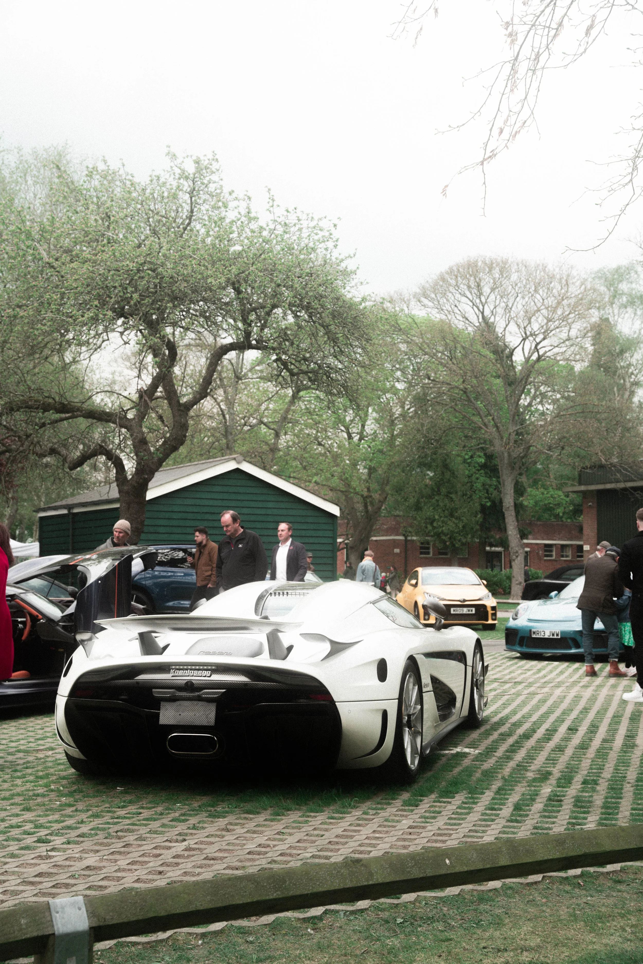 A white sports car with a rear wing and sleek design parked on a patterned brick surface at an outdoor car show, with several people and other cars in the background, trees, and a green shed.