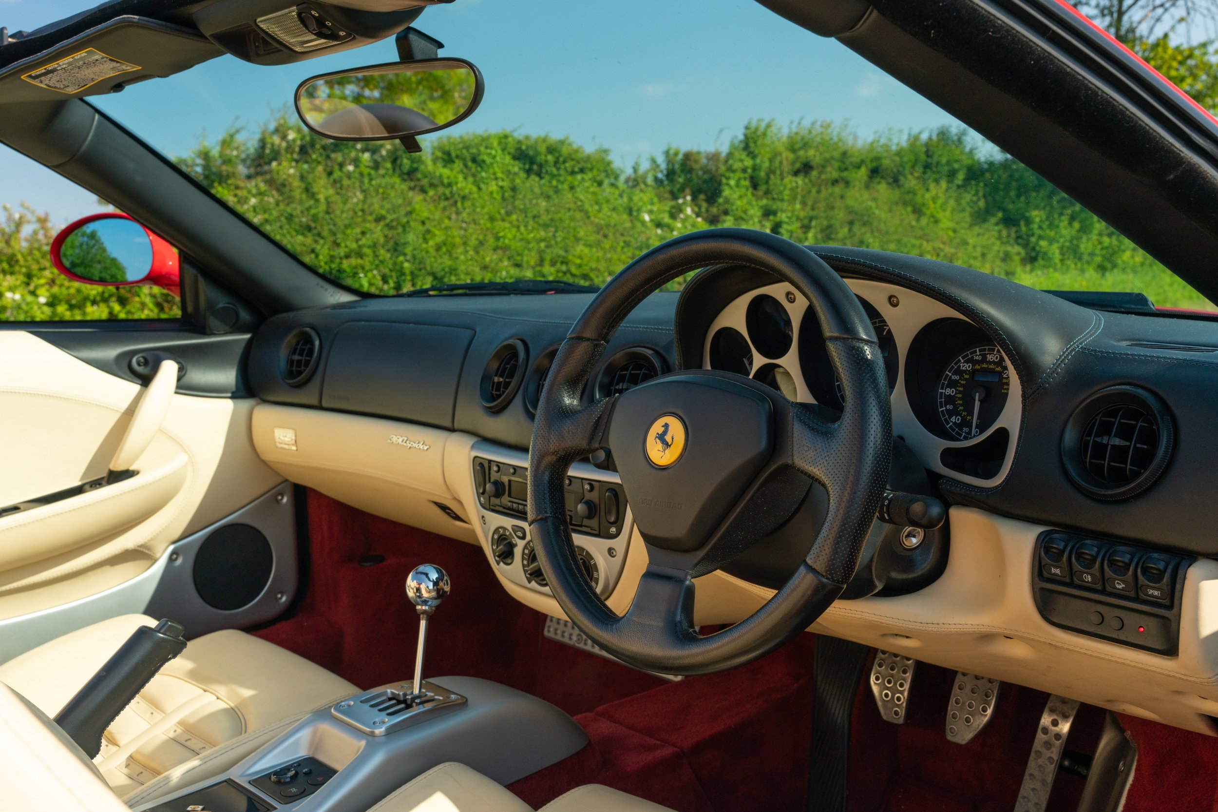 Interior of a Ferrari sports car with a black steering wheel featuring the Ferrari emblem, black and beige dashboard, manual gear shift, and red carpeting, with green foliage visible outside the windshield.