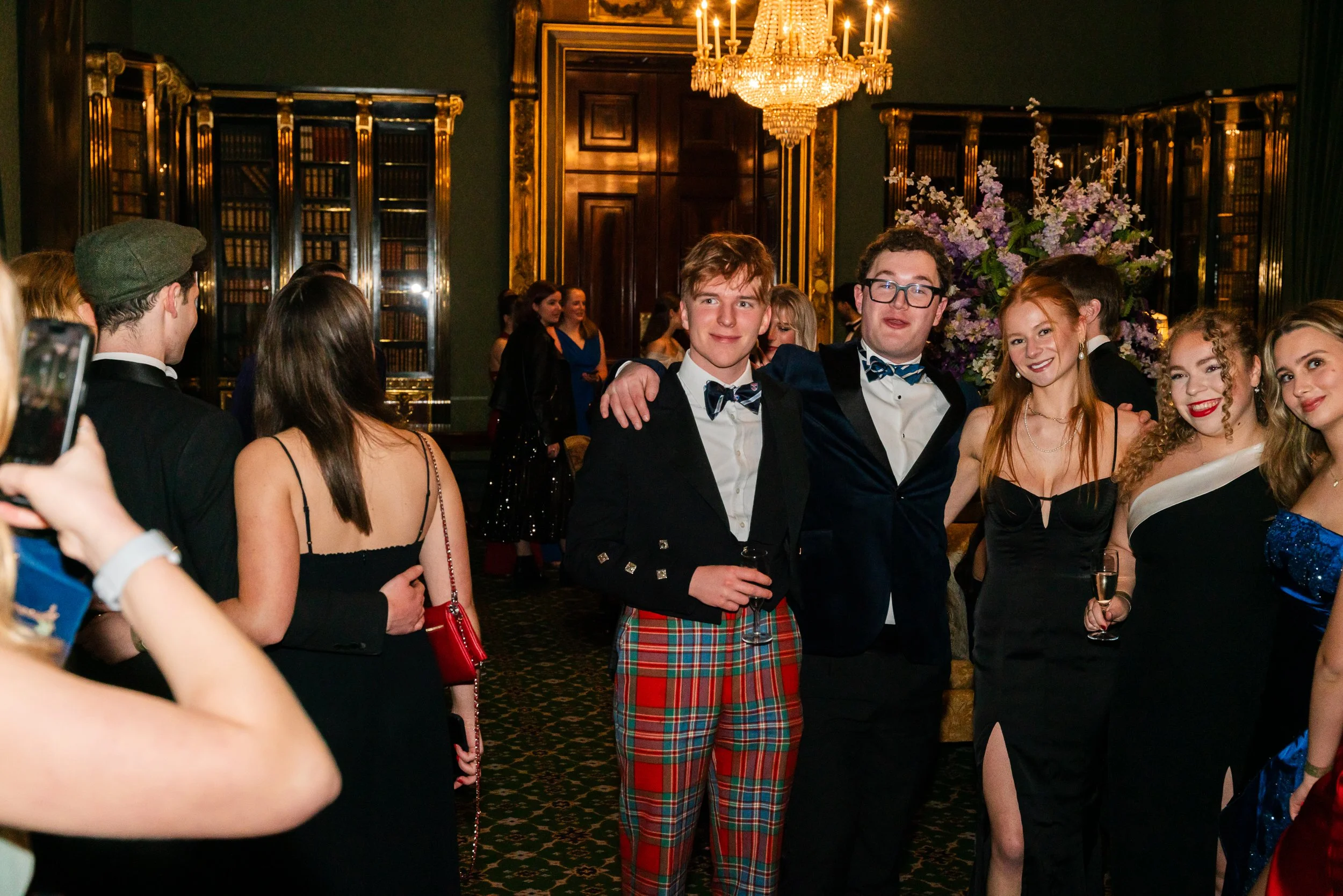 Group of young adults dressed in formal attire at an elegant event, posing for photo in a luxurious room with chandeliers, dark green walls, gold accents, and bookshelves.