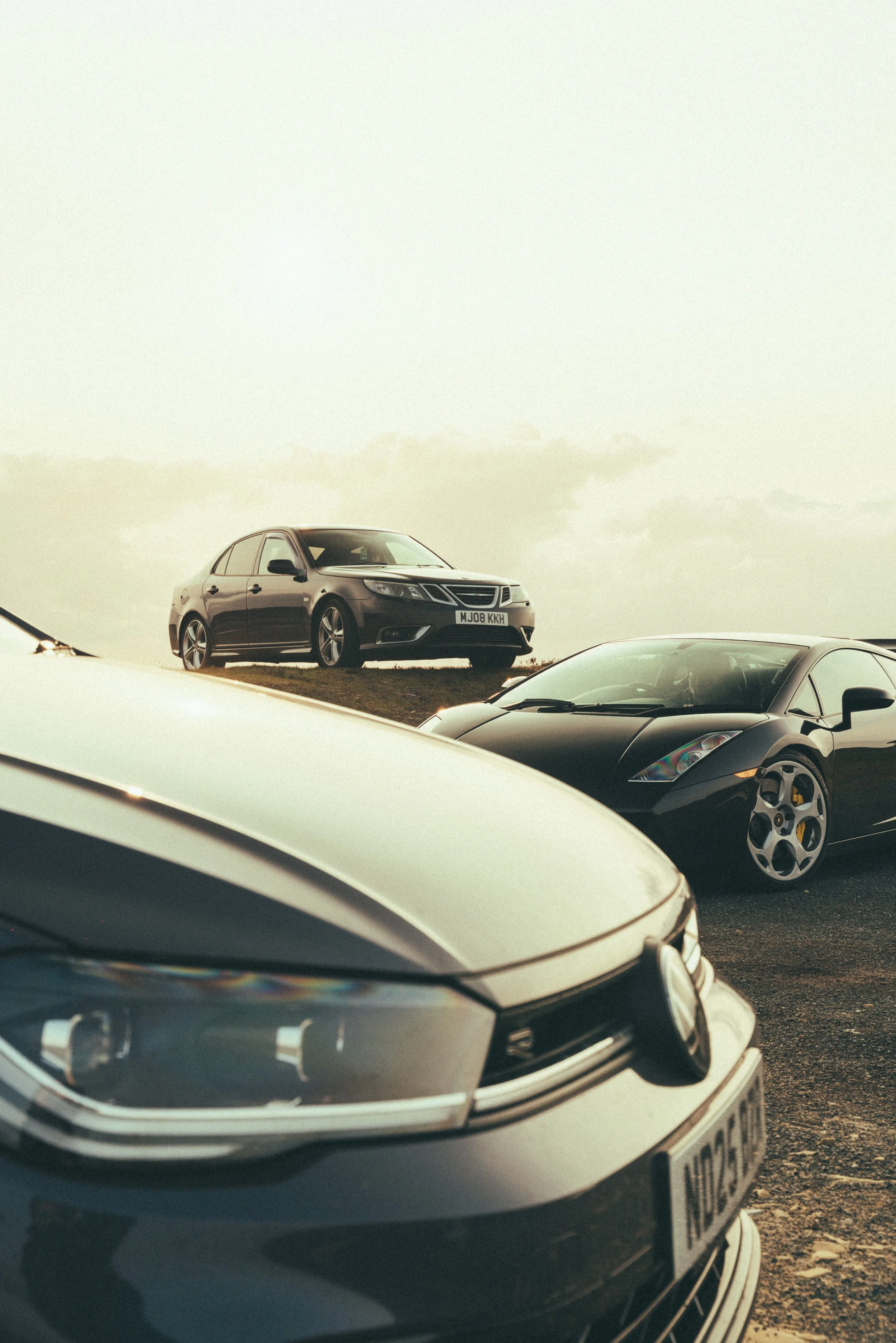 Multiple luxury cars parked outdoors with a backdrop of a cloudy sky, including a close-up of a white car in the foreground, a black Lamborghini to the right, and a silver Saab on a hill in the background.