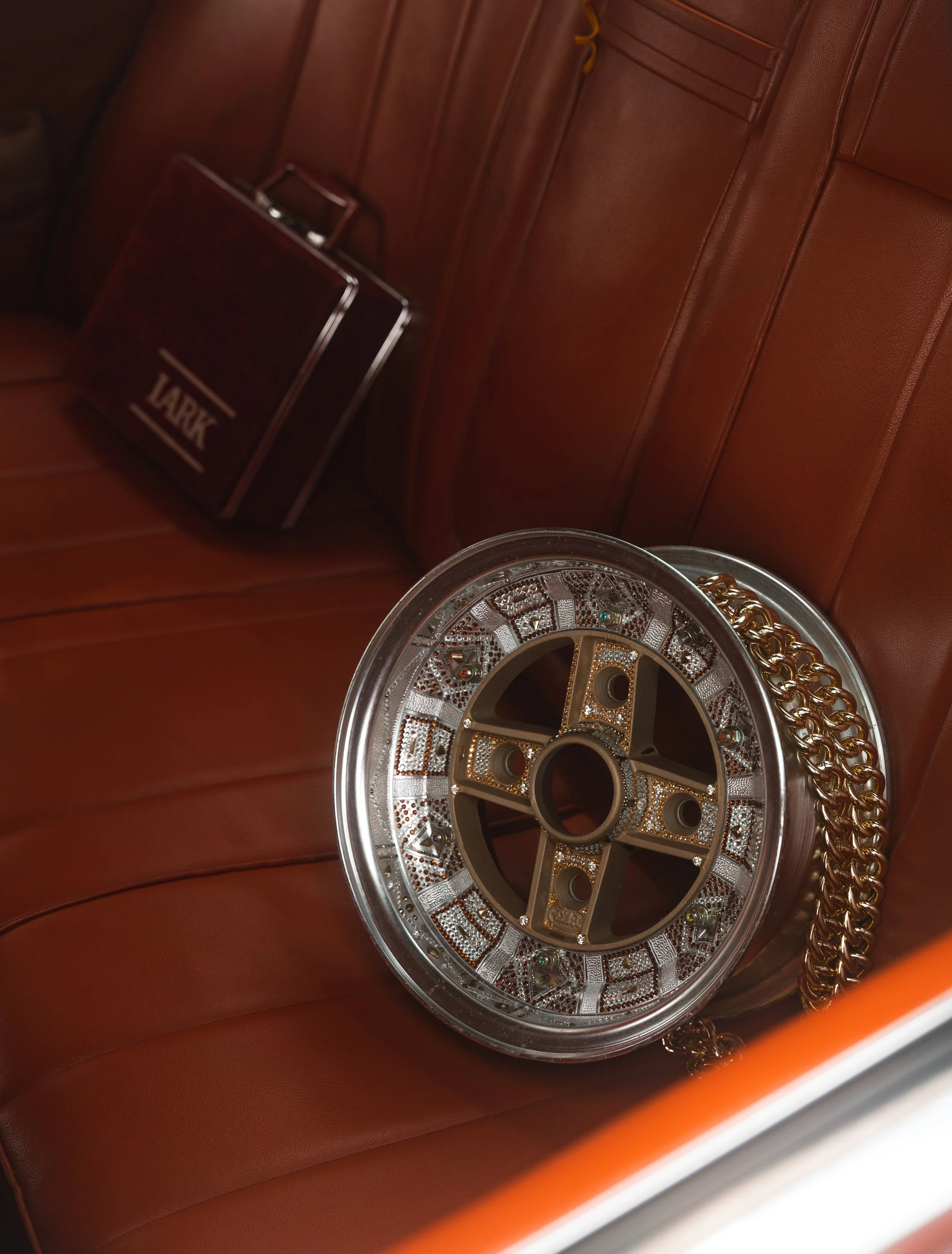 A shiny jewelry wheel with gold accents, chains, and embedded gemstones on a reddish-brown leather car seat, with a briefcase in the background.