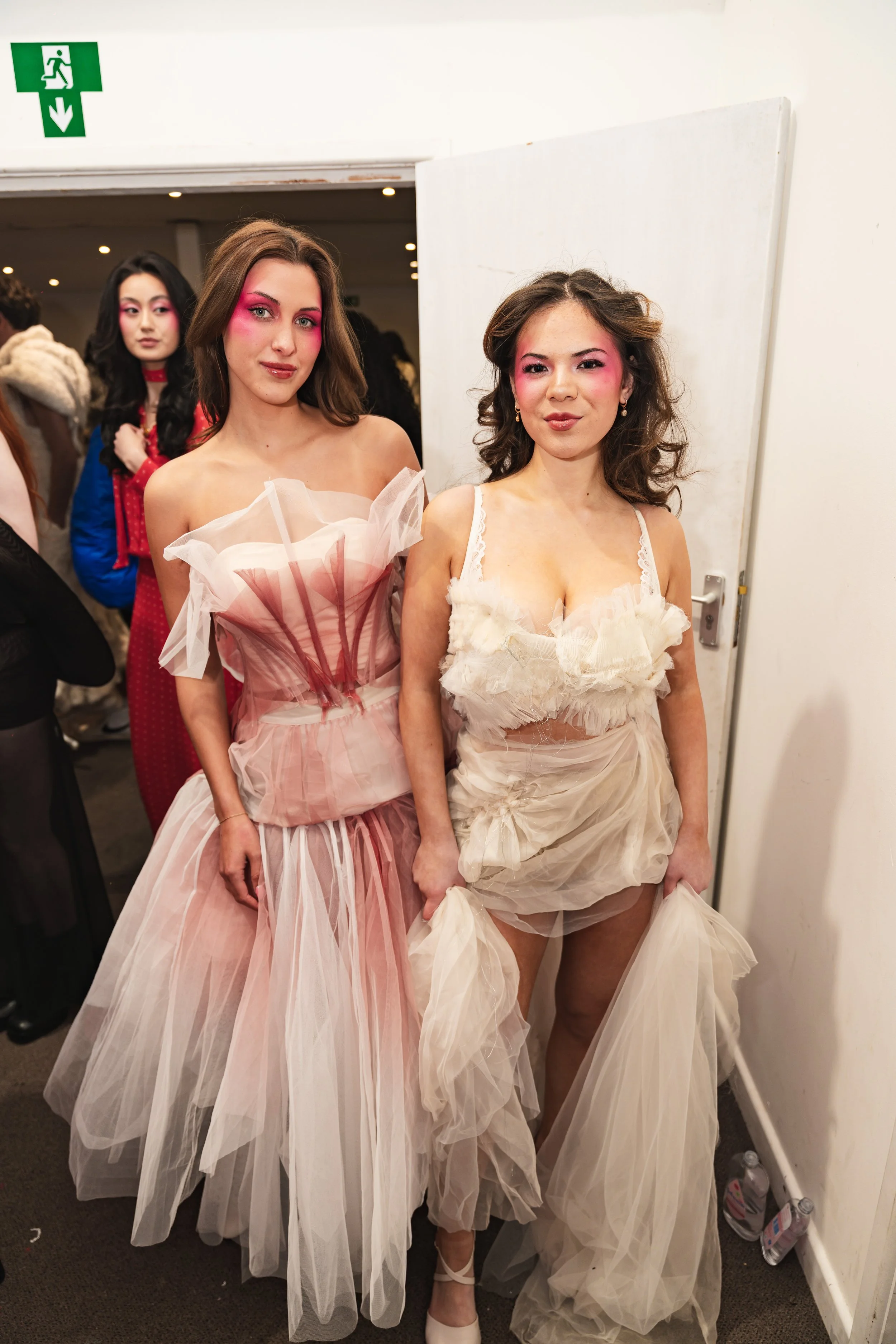 Two women wearing elaborate tulle dresses with pink and cream color tones, makeup featuring bold pink eyeshadow, standing back-to-back, in a room with other people and emergency exit sign overhead.